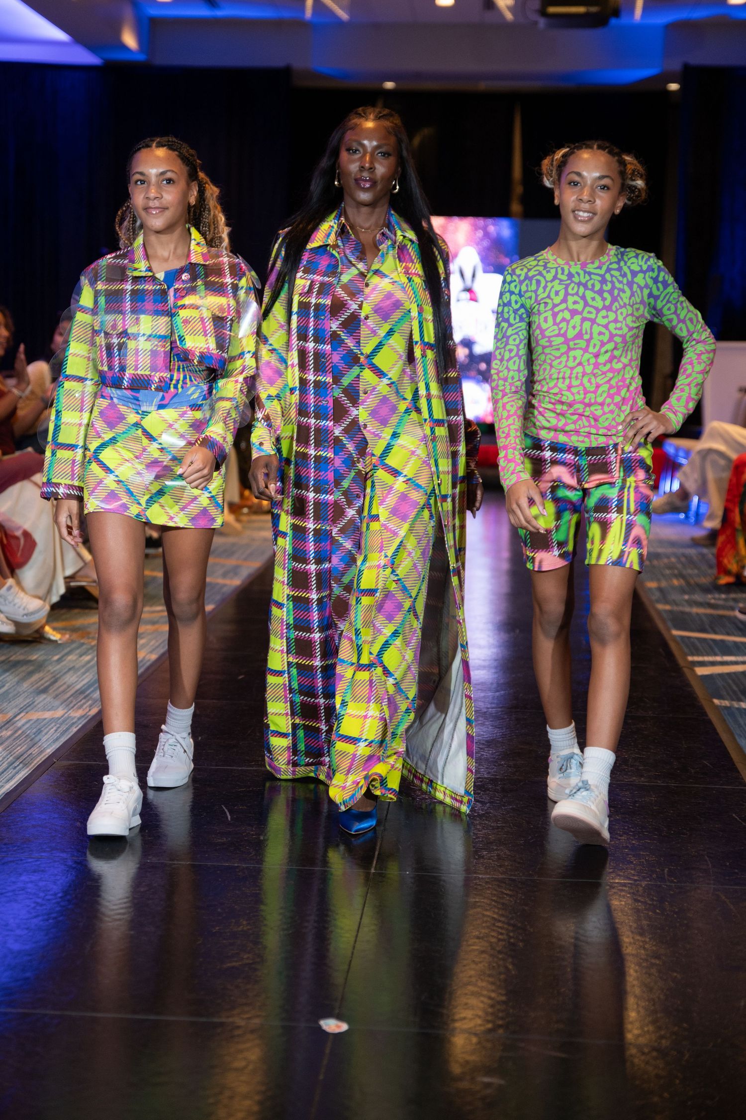 Three models walk the runway in vibrant neon plaid and animal print outfits at a colorful fashion show.