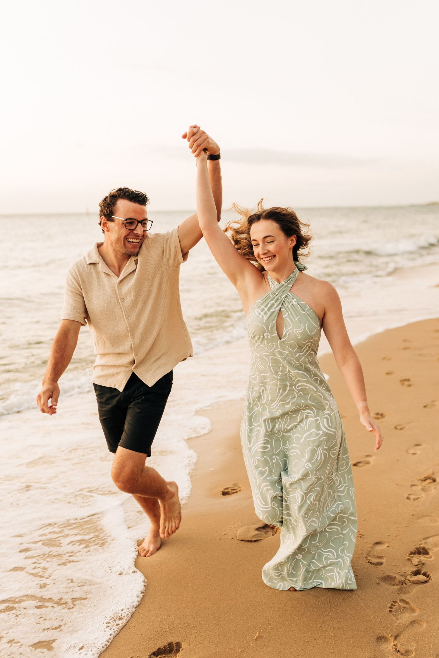 Joyful couple holding hands and running along beach shoreline at sunset while wearing casual summer attire.