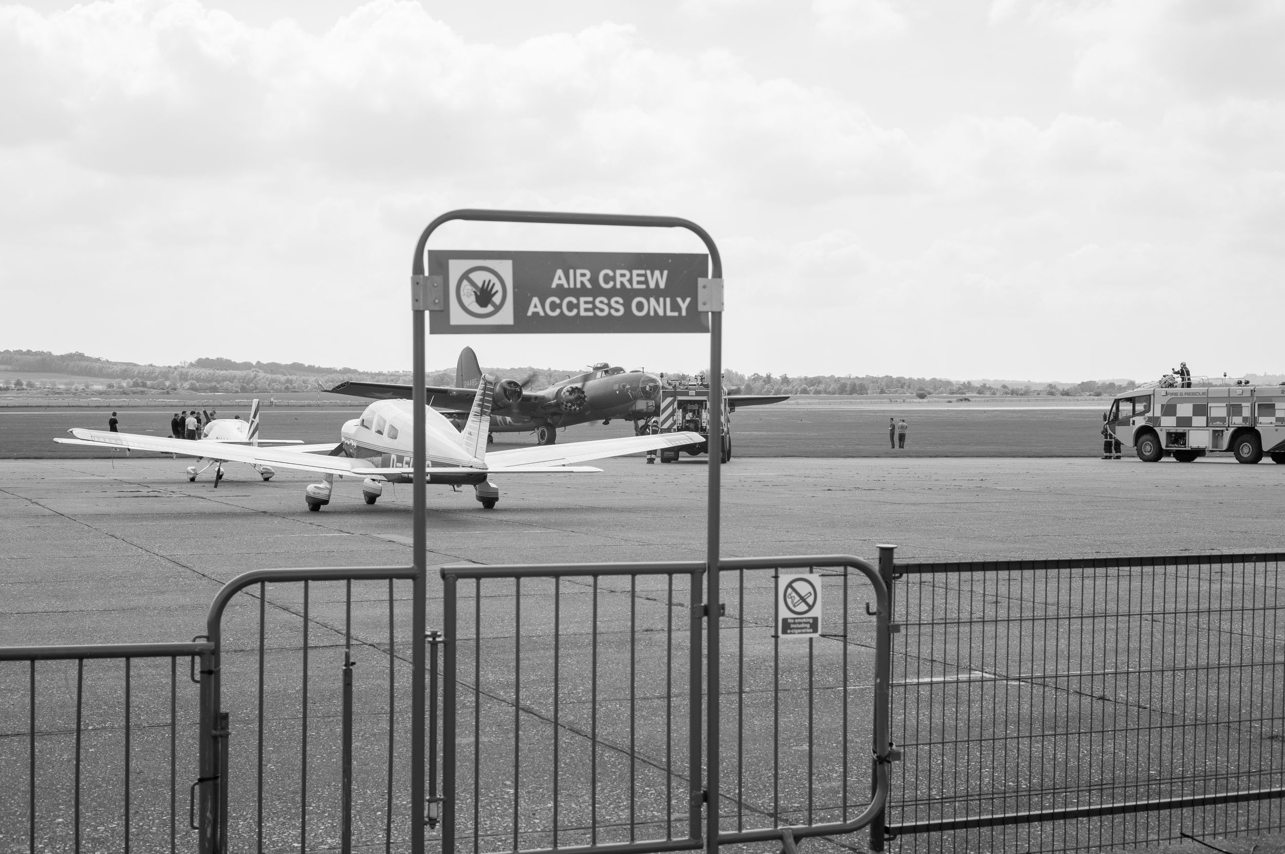 Black and white photo of an airfield with Air Crew Access Only sign, vintage aircraft, and ground vehicles on the tarmac.