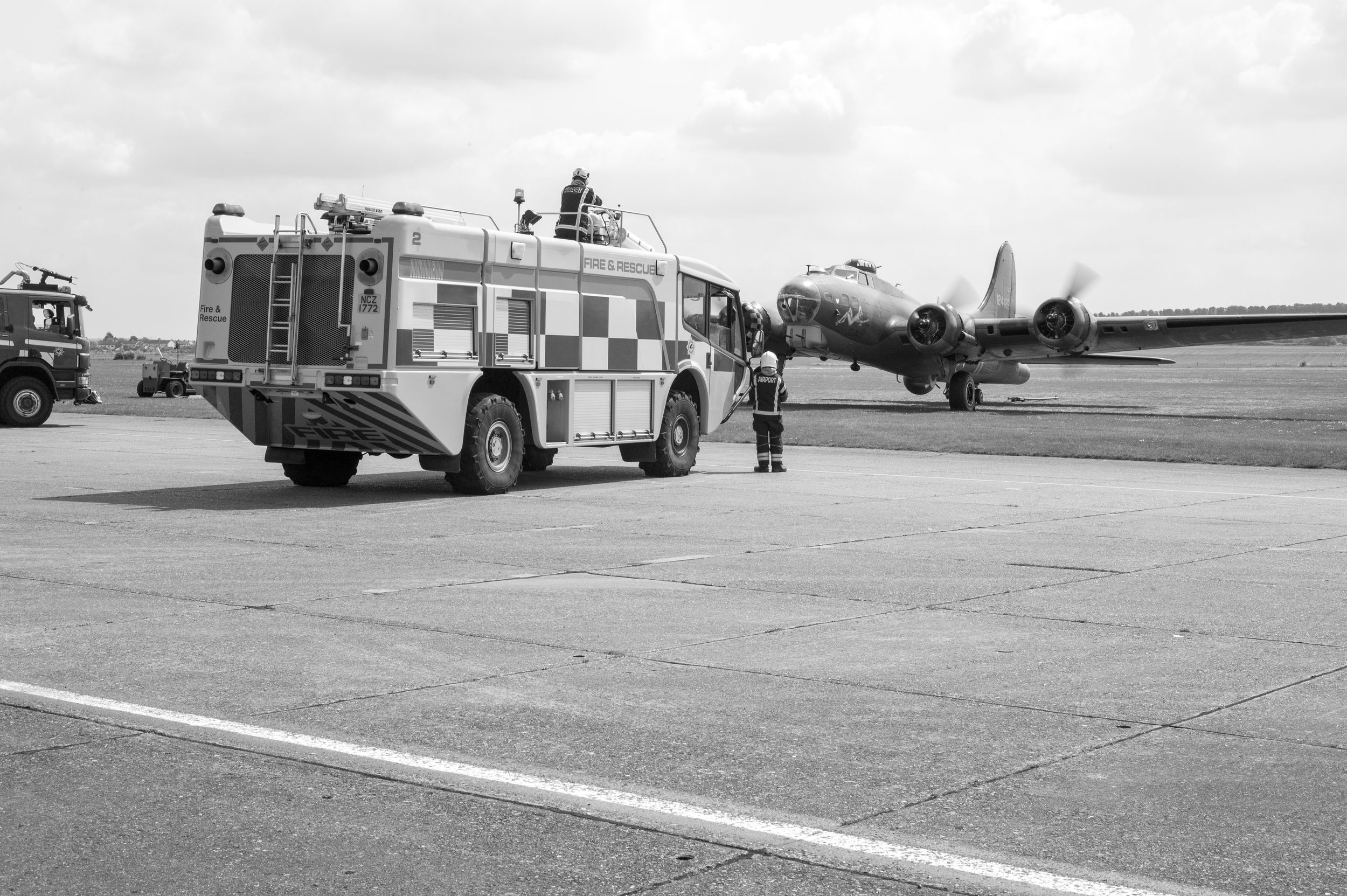 Black and white photo of airport fire truck and personnel on tarmac with vintage propeller aircraft in background.