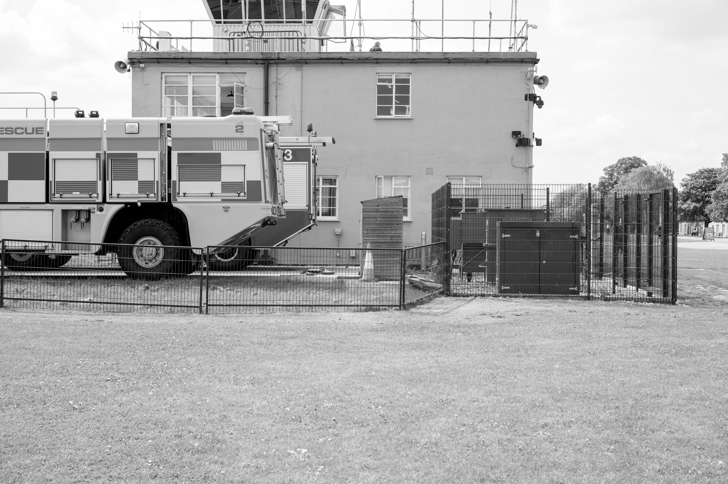 Black and white photo of a fire truck parked beside an airport control tower behind a metal fence.