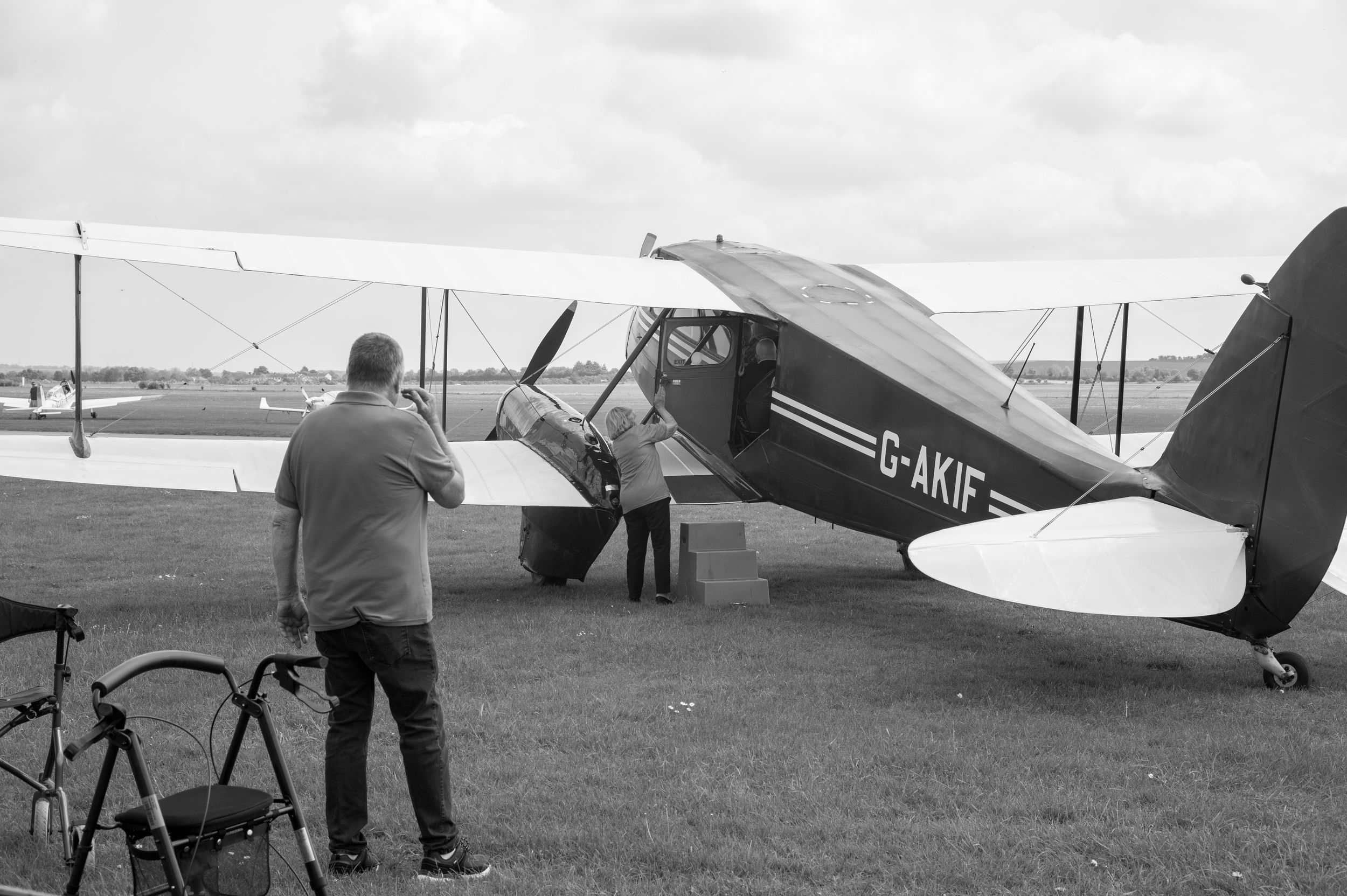 Two men examine a vintage biplane marked G-AKIF on a grass airfield in this classic black and white photograph.
