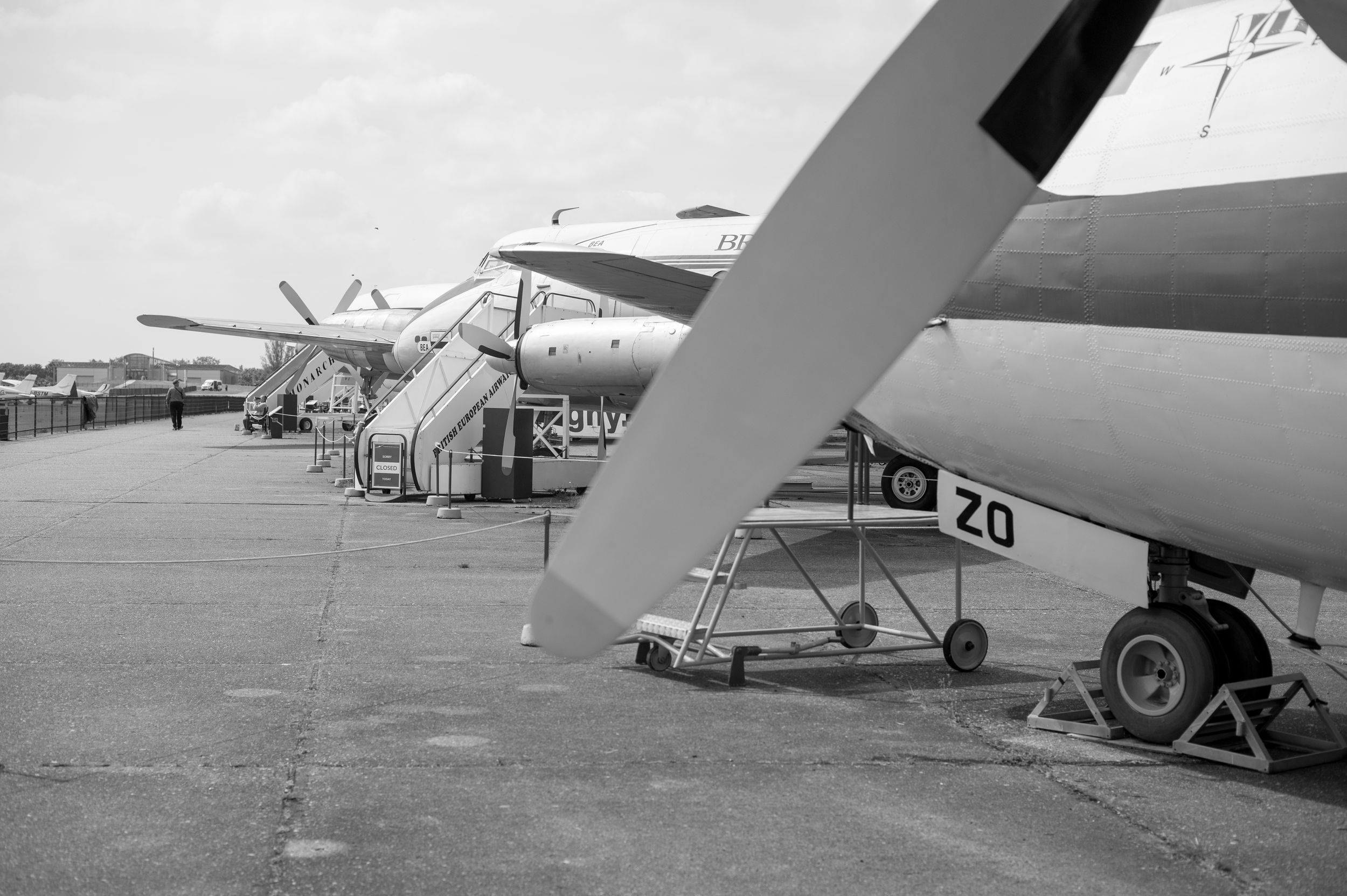 Black and white photo of multiple vintage propeller airplanes lined up on an airport tarmac under a cloudy sky.
