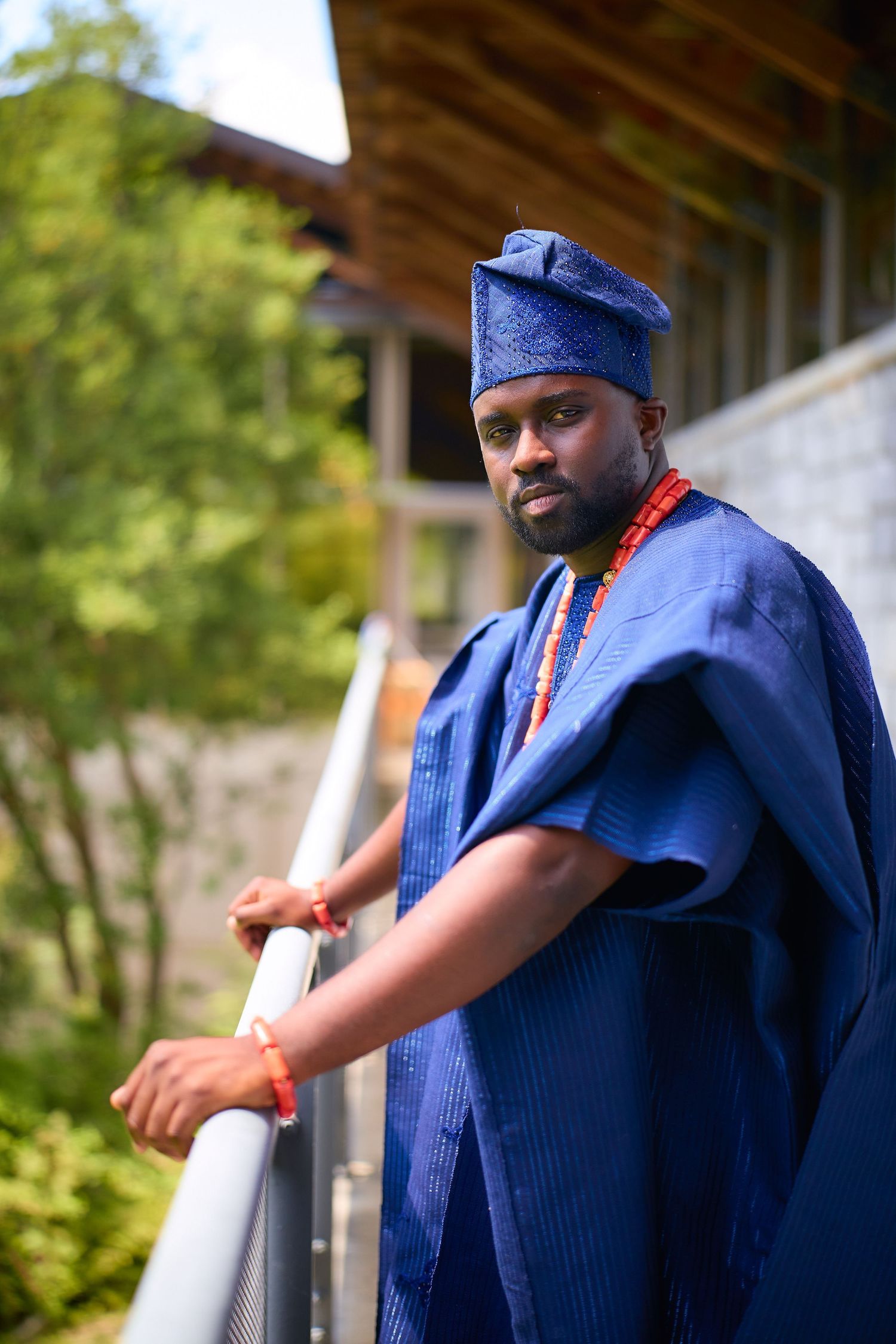 Man in blue traditional Nigerian Agbada attire and cap standing on a balcony surrounded by greenery.