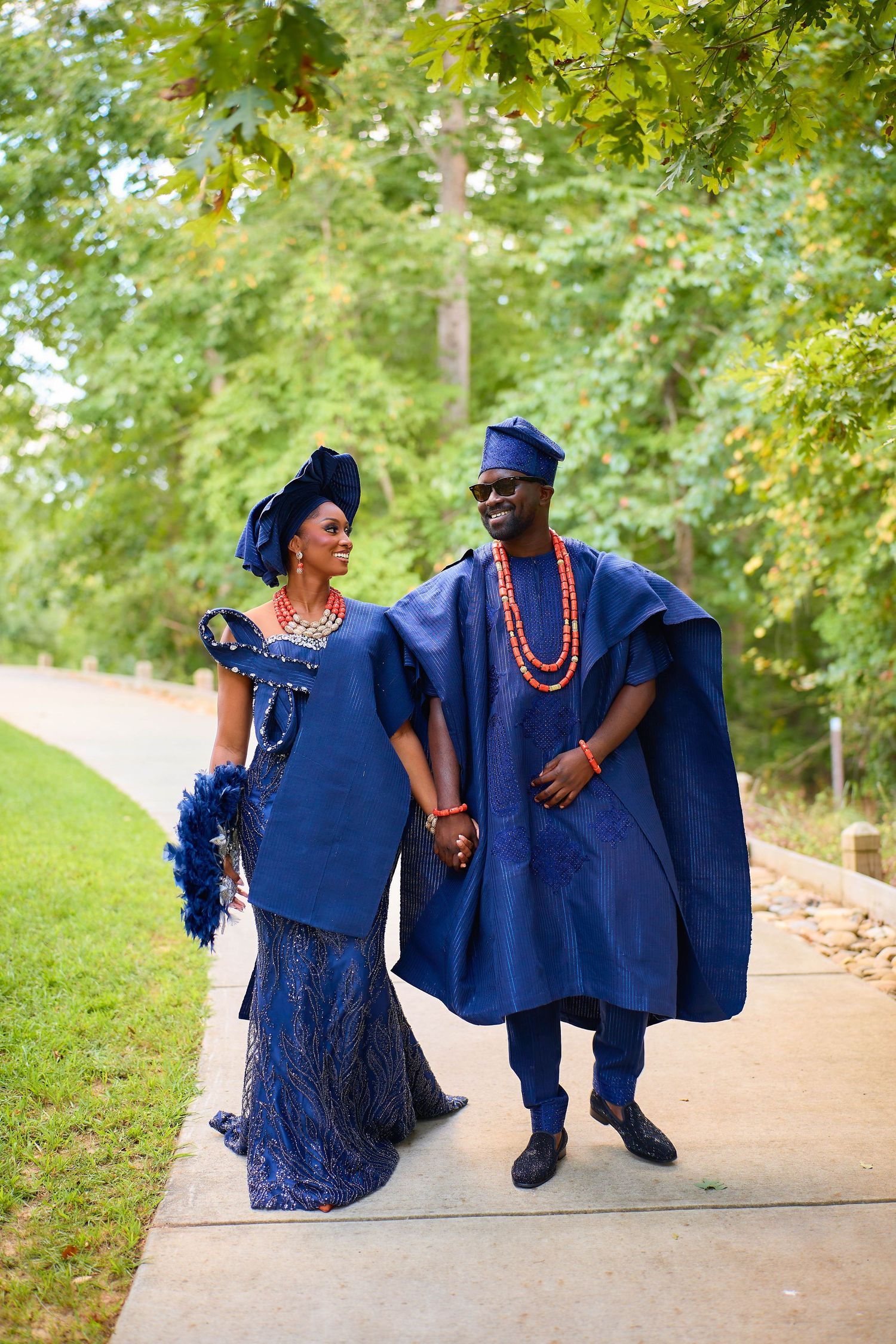 Couple in matching navy blue traditional African attire walking hand in hand on a path surrounded by lush green trees.