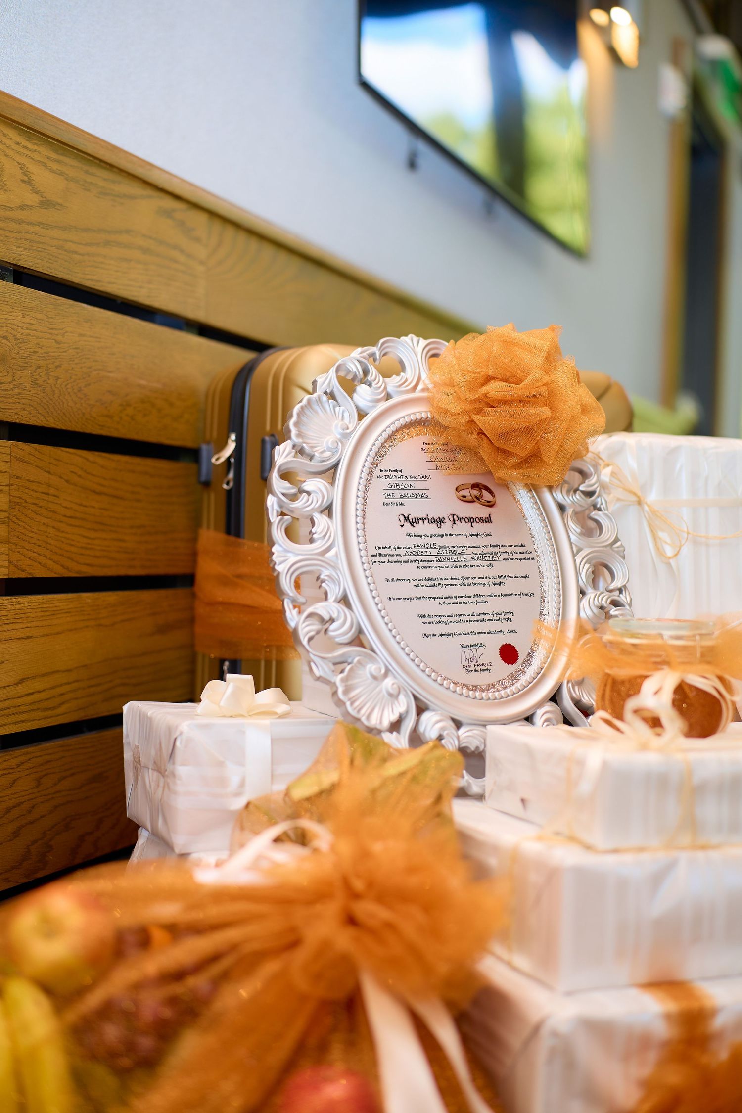 Elegant wedding proposal display with ornate white frame, gifts wrapped in white, and orange decorative bows on a wooden bench.