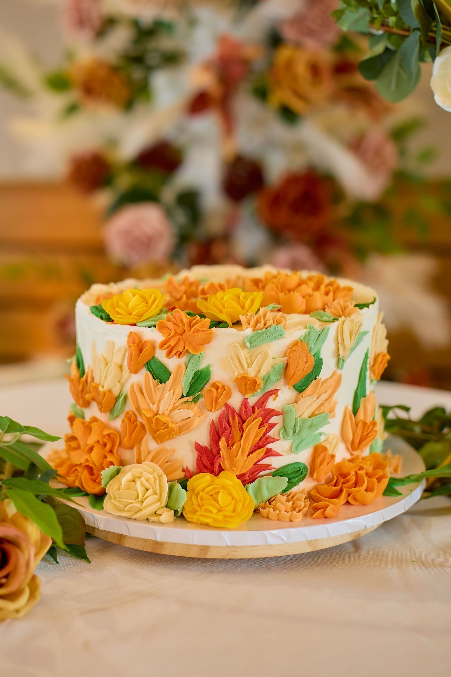 Elegant fall wedding cake with orange, yellow, and green buttercream floral decorations on white frosting, displayed on a table.