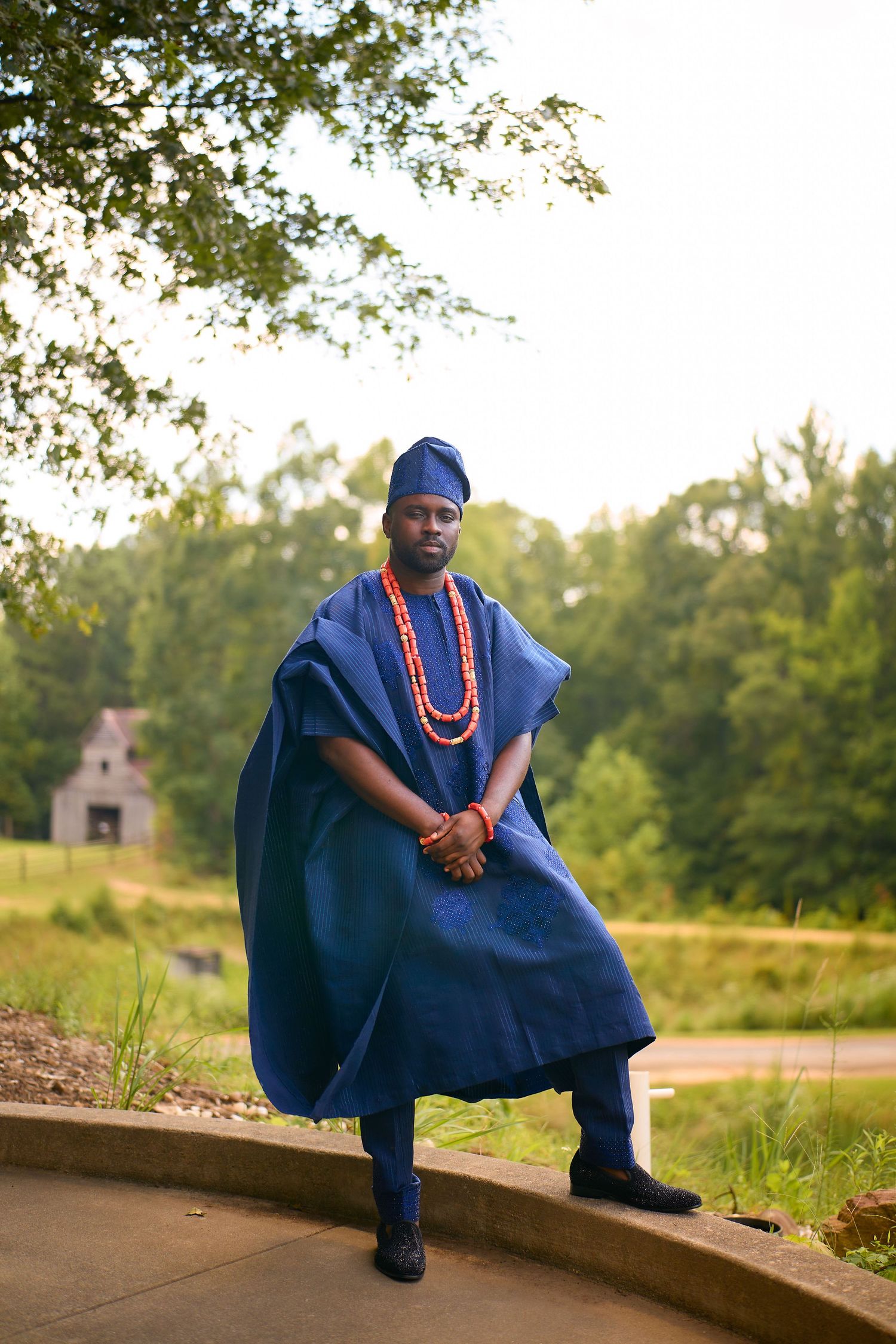 Man in traditional blue Nigerian Agbada attire with red coral beads standing outdoors in a lush green setting.