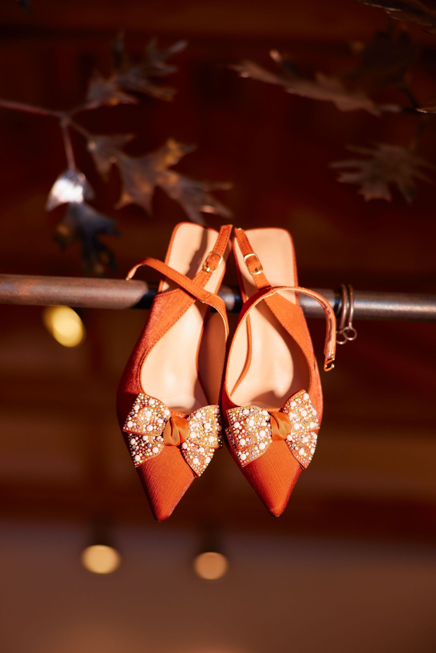 Orange satin pointed-toe flats with rhinestone bow embellishments hanging from a rustic metal rod with decorative leaves.