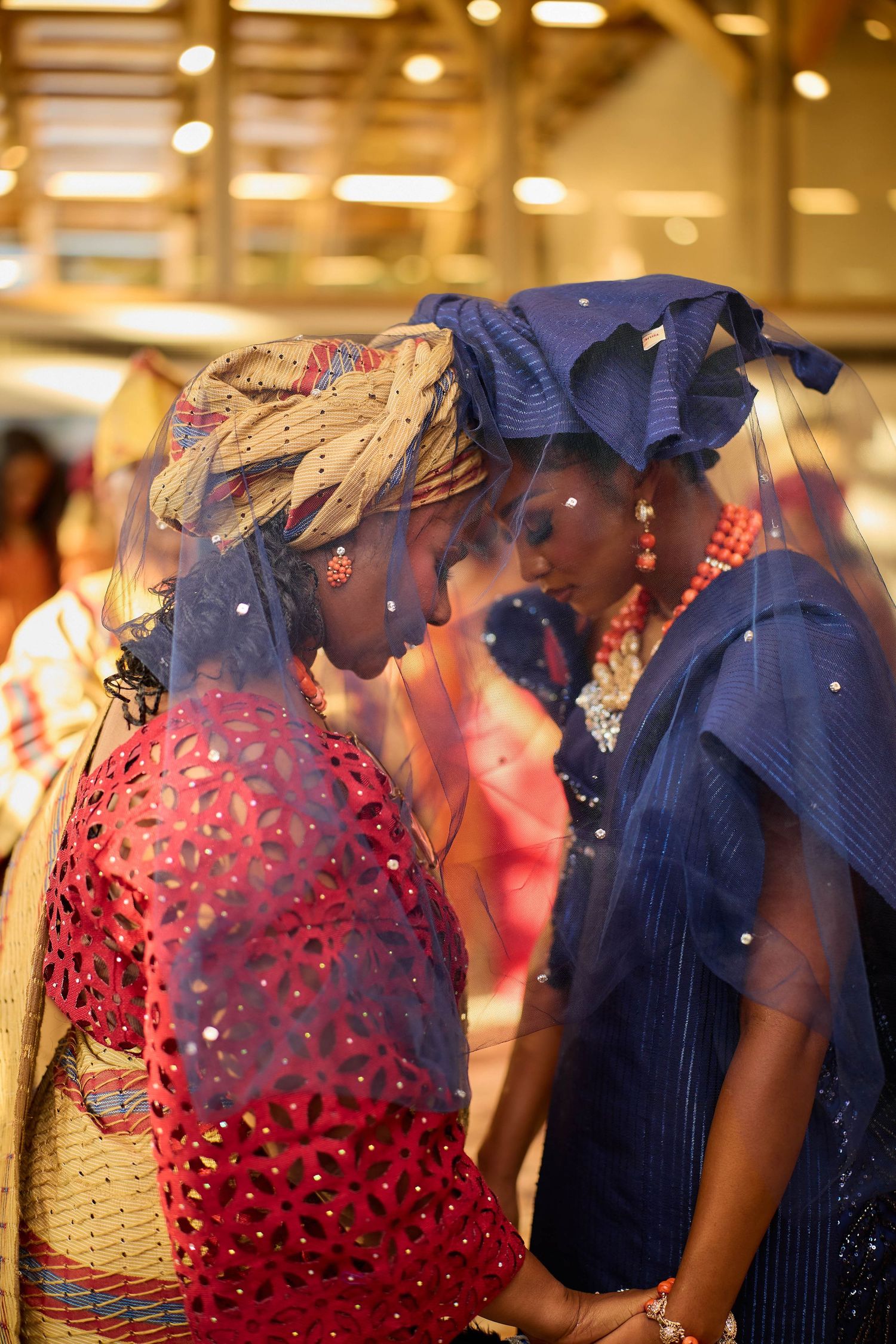 Two women in vibrant traditional African attire and head wraps share an intimate moment under a sheer blue veil.