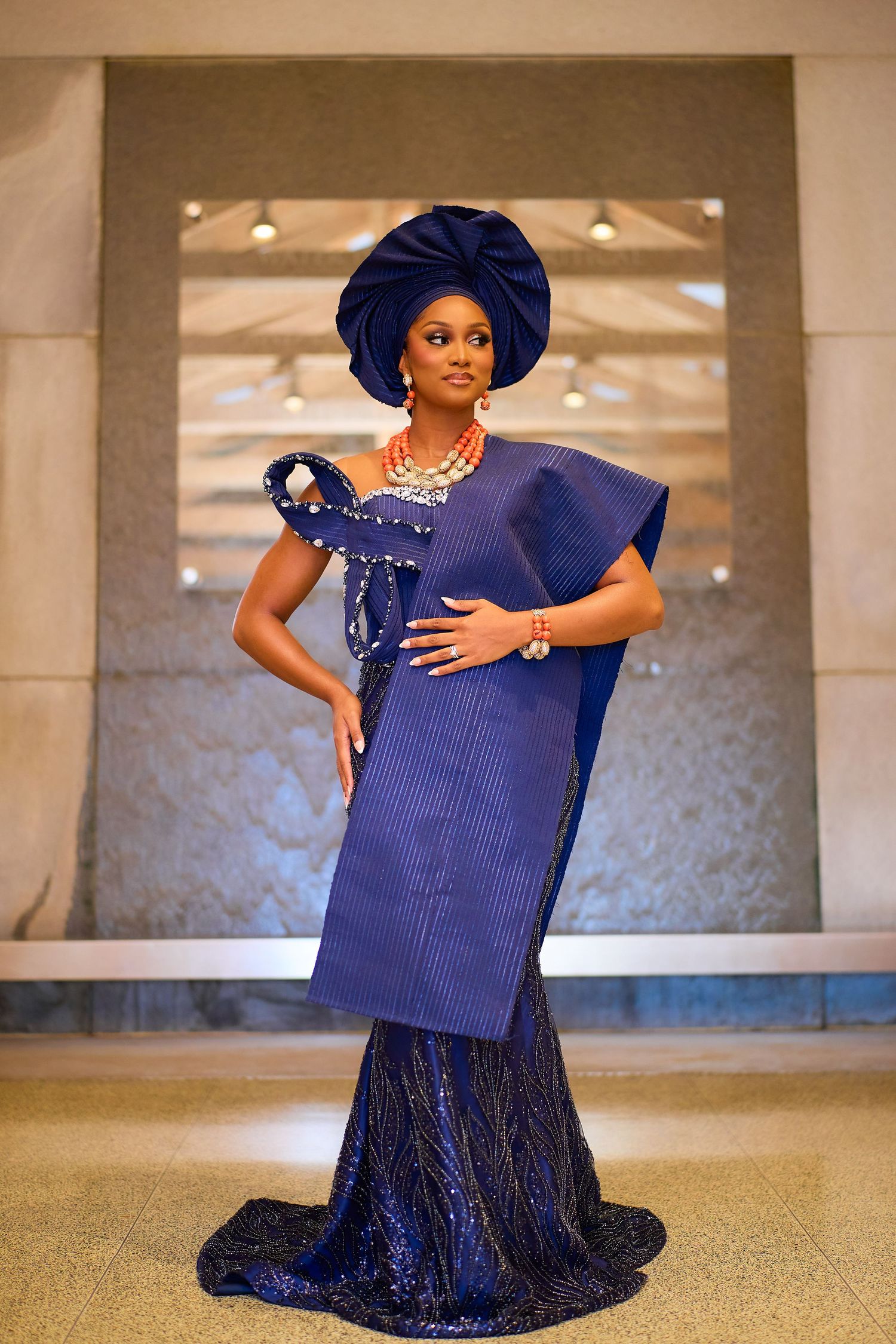 Woman in elegant navy blue Nigerian traditional attire with gele headwrap and coral beaded jewelry.
