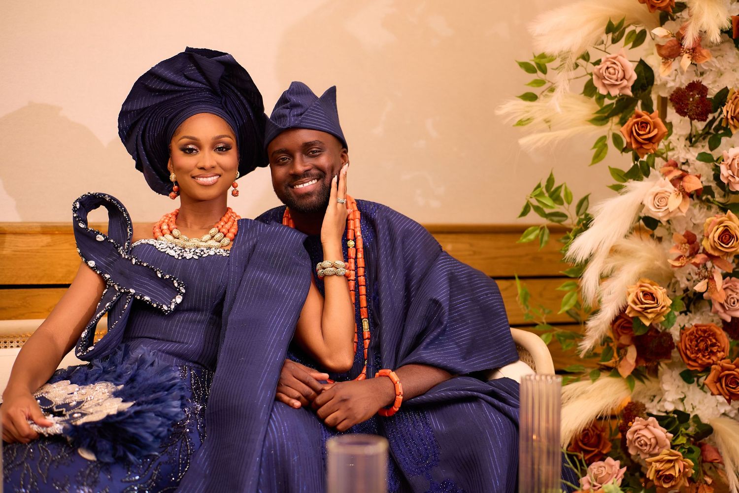 Nigerian couple in matching navy blue traditional attire with coral beads, seated by floral arrangement.