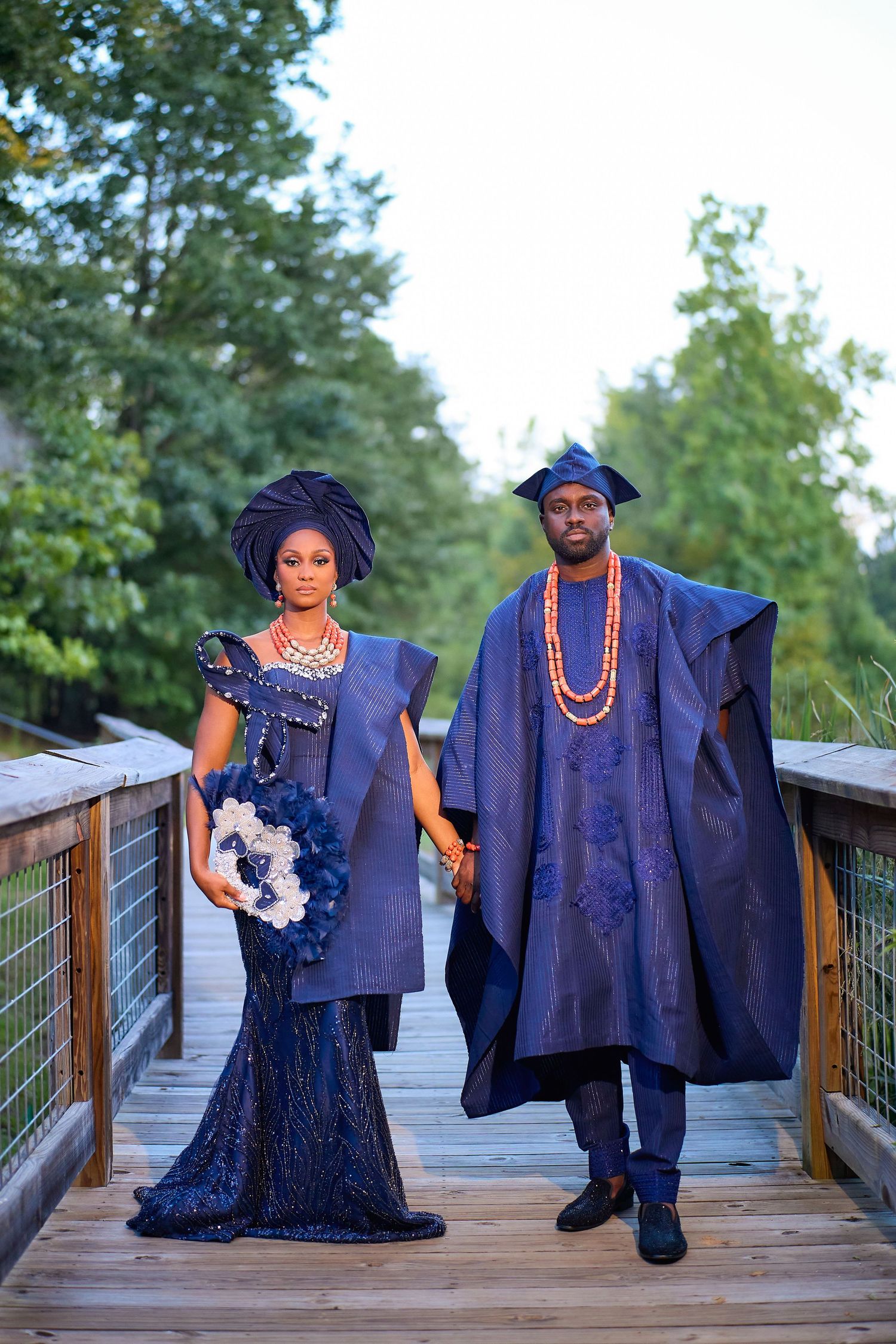 Couple in matching navy blue traditional African attire posing on a wooden bridge surrounded by lush green trees.