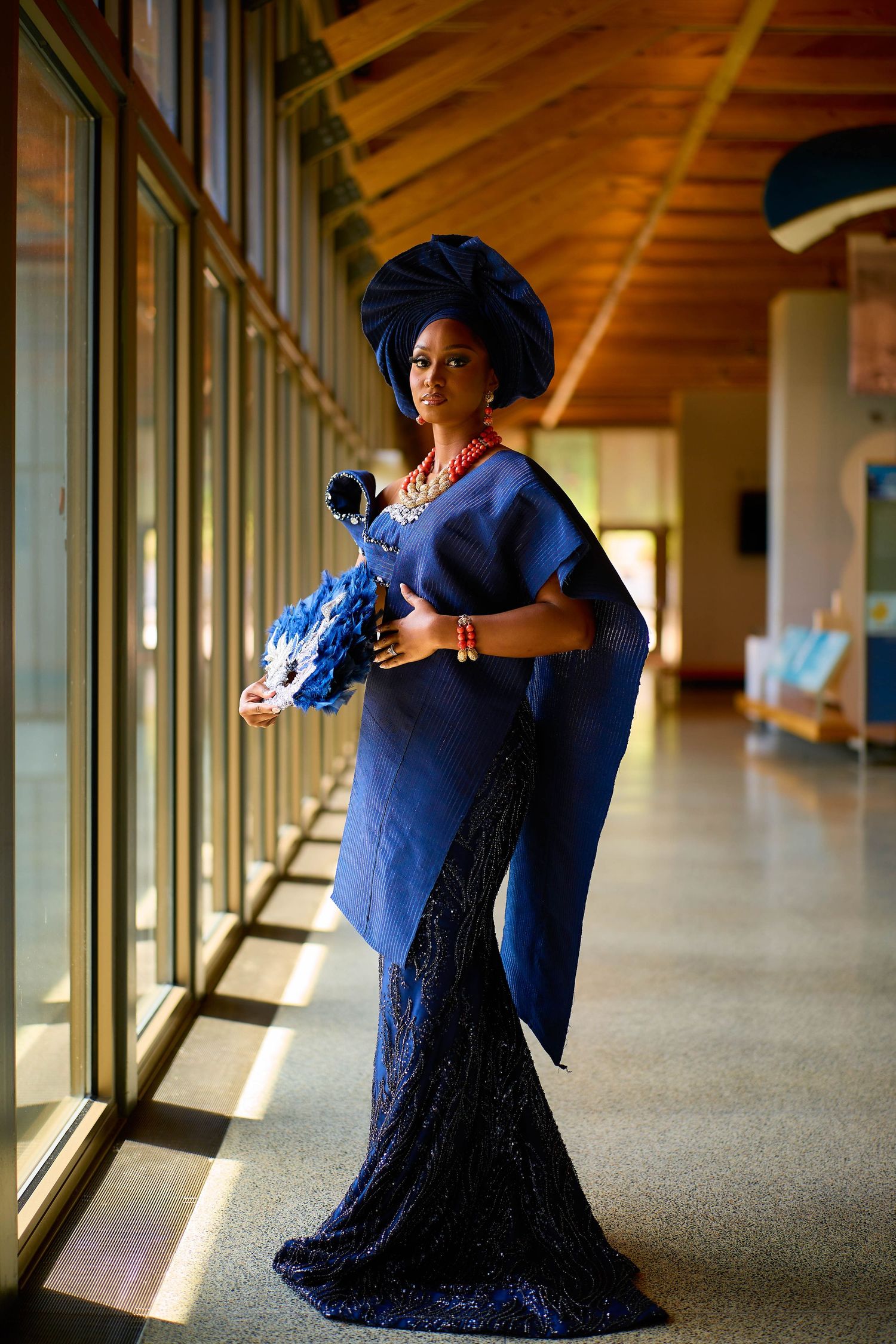 Woman in elegant blue Nigerian traditional attire with gele headwrap and coral beads poses by windows.