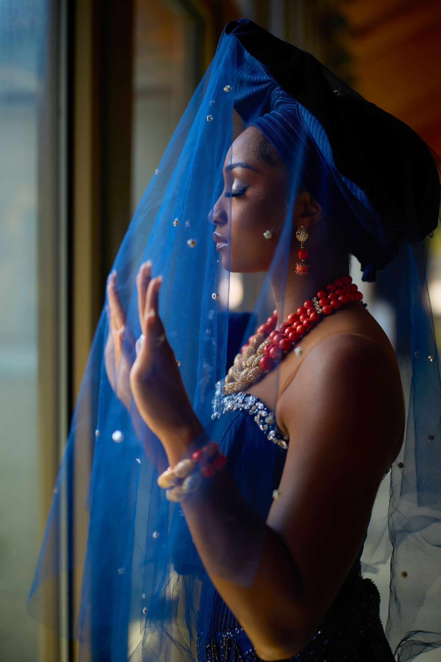 A Black woman in blue traditional attire with red coral beads and veil poses gracefully by a window.