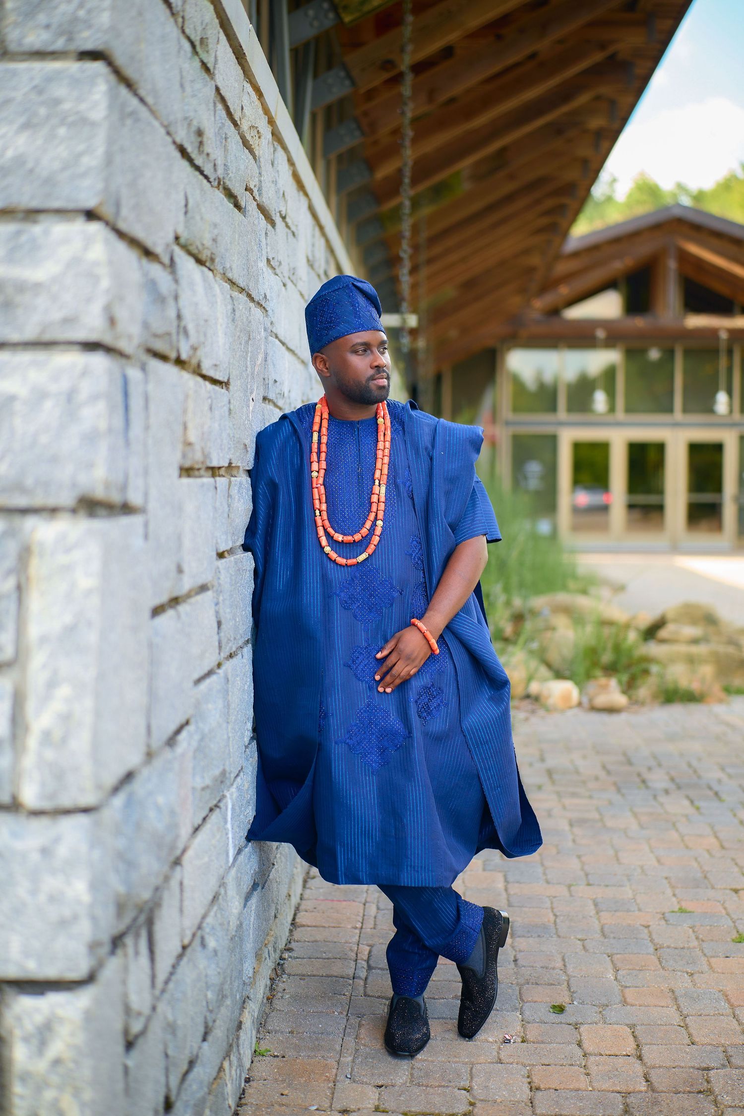 Man in blue Nigerian agbada traditional attire with red coral beads necklace leaning against stone wall outdoors.
