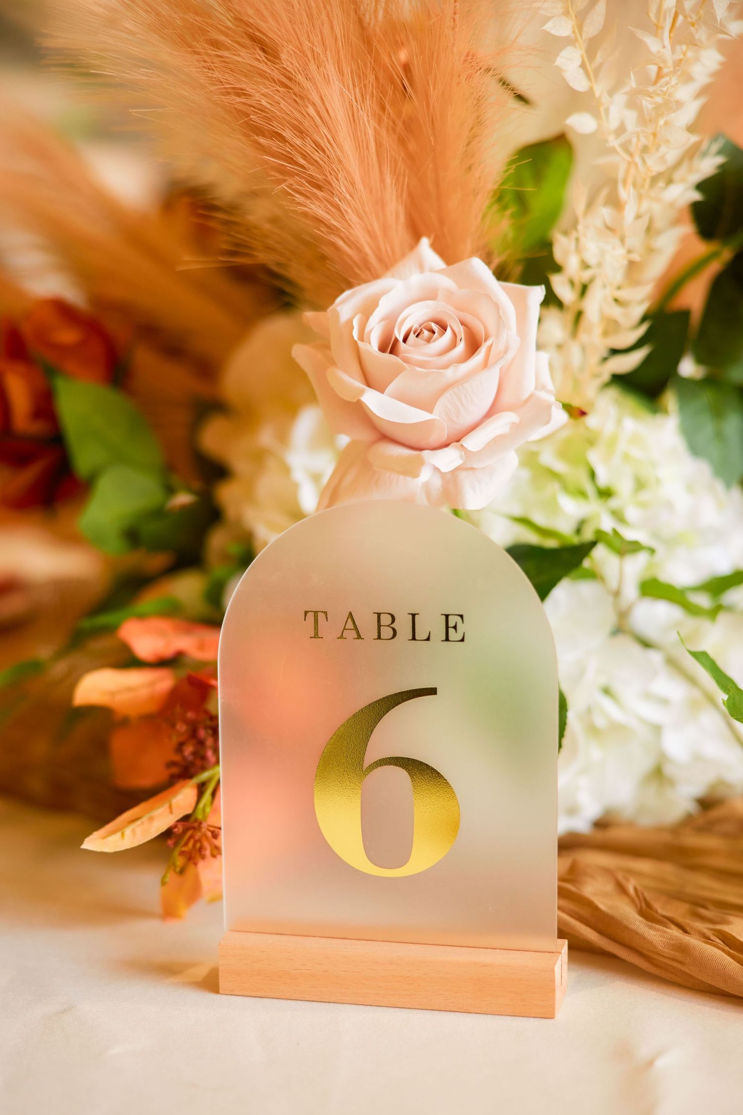 Elegant wedding table number 6 sign on frosted acrylic with gold text, pink rose, and boho floral arrangement.