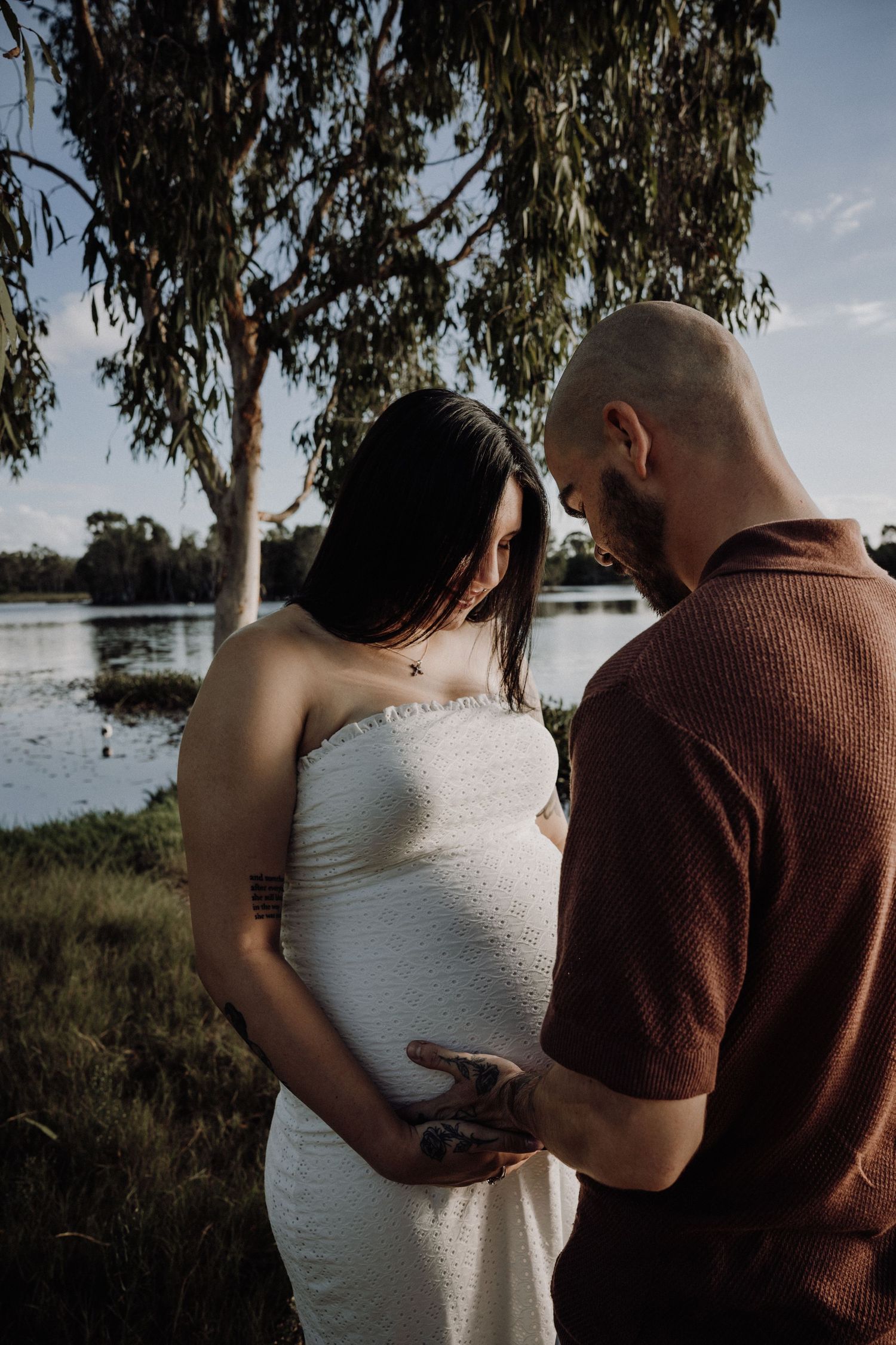 Couple tenderly cradling pregnant belly outdoors by a serene lake, surrounded by lush greenery at golden hour.