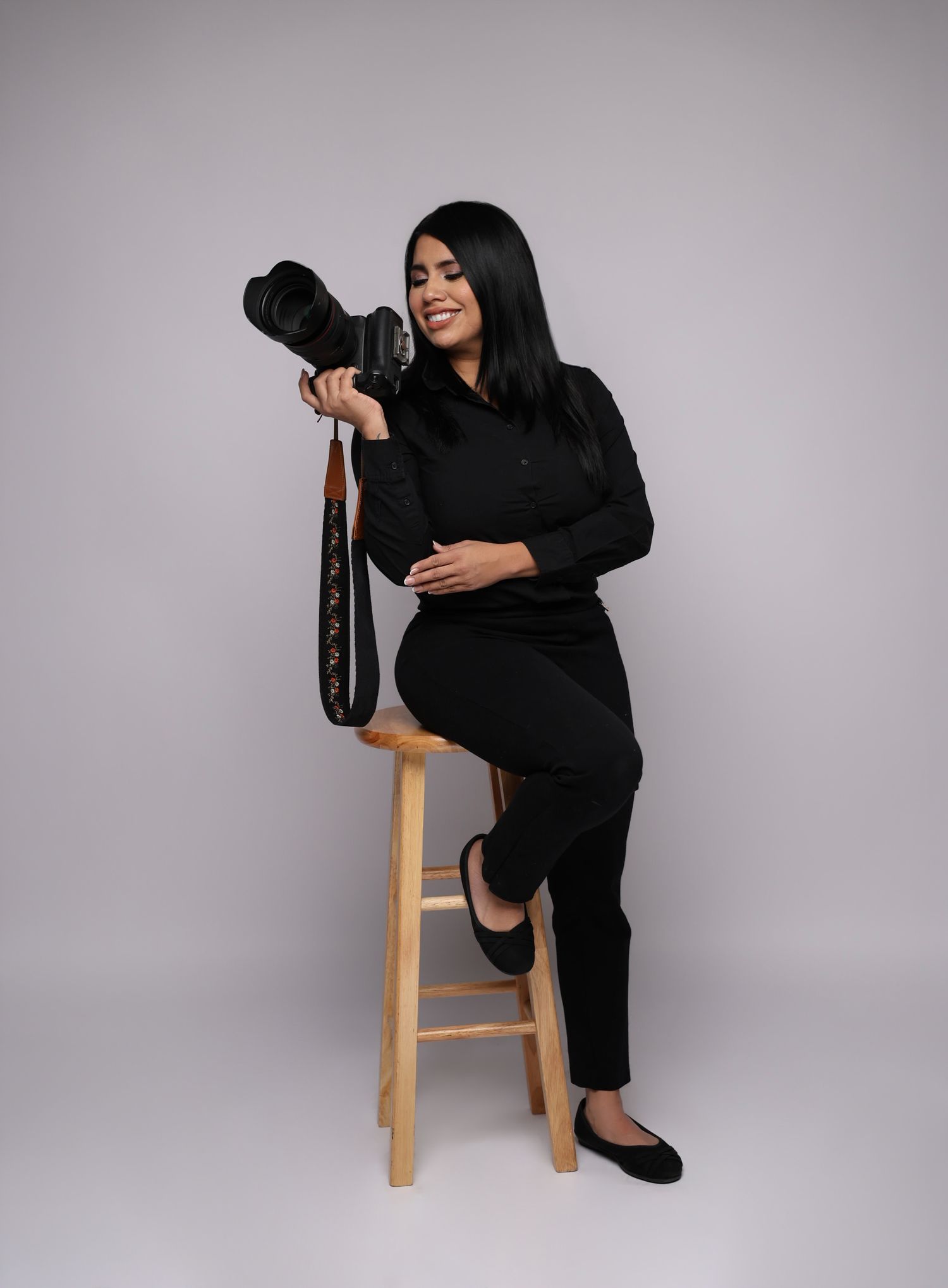 Woman in black outfit holding a professional DSLR camera against a brown studio background.