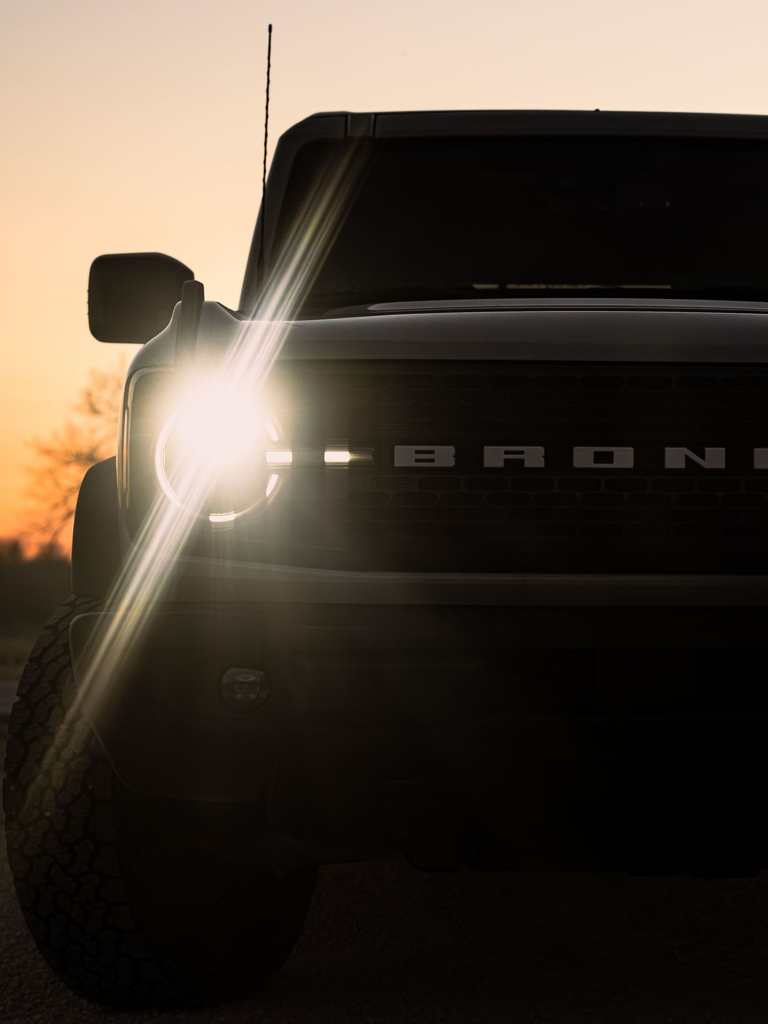Ford Bronco SUV silhouetted against a golden sunset with bright headlight flare in dramatic low-light photography.