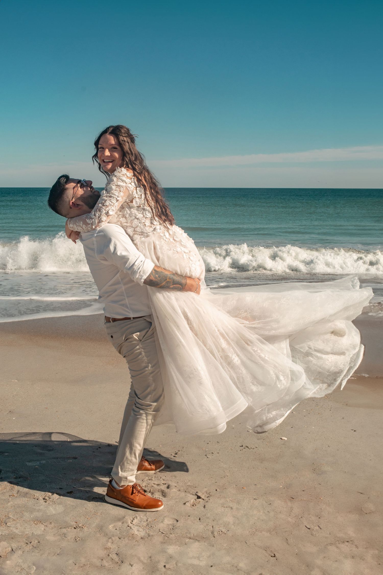 Beach wedding couple in dramatic pose with flowing white dress against ocean waves.