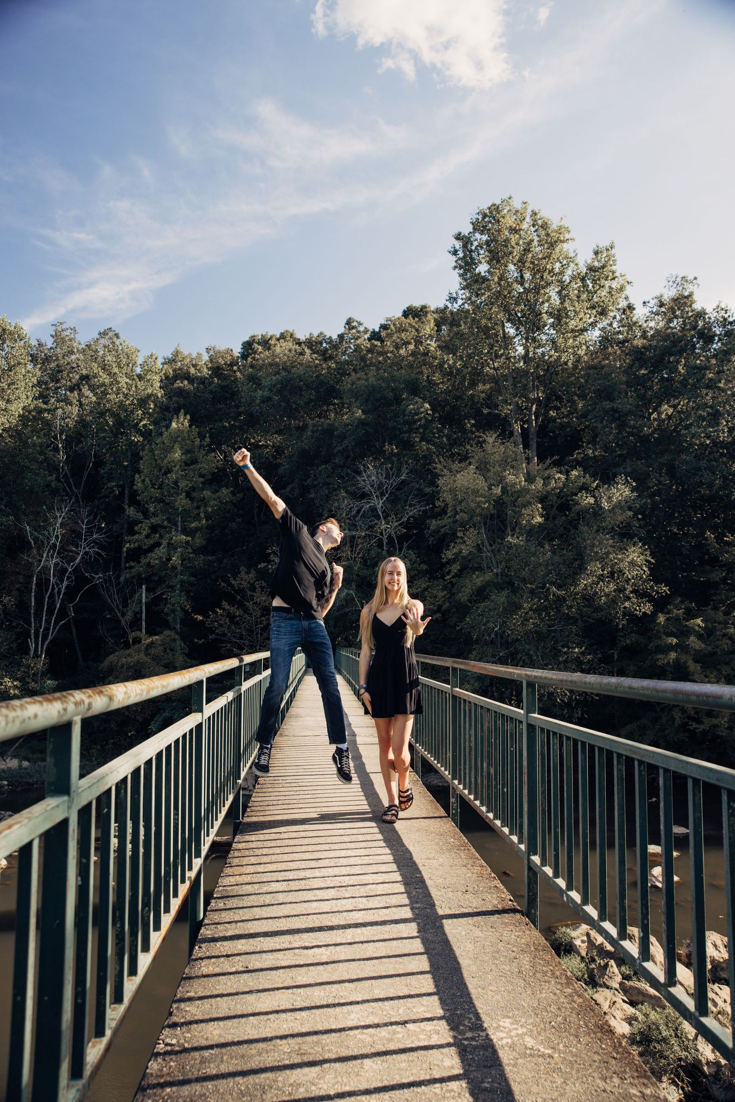 Two people enjoying a sunny day while standing on a metal footbridge surrounded by green trees.