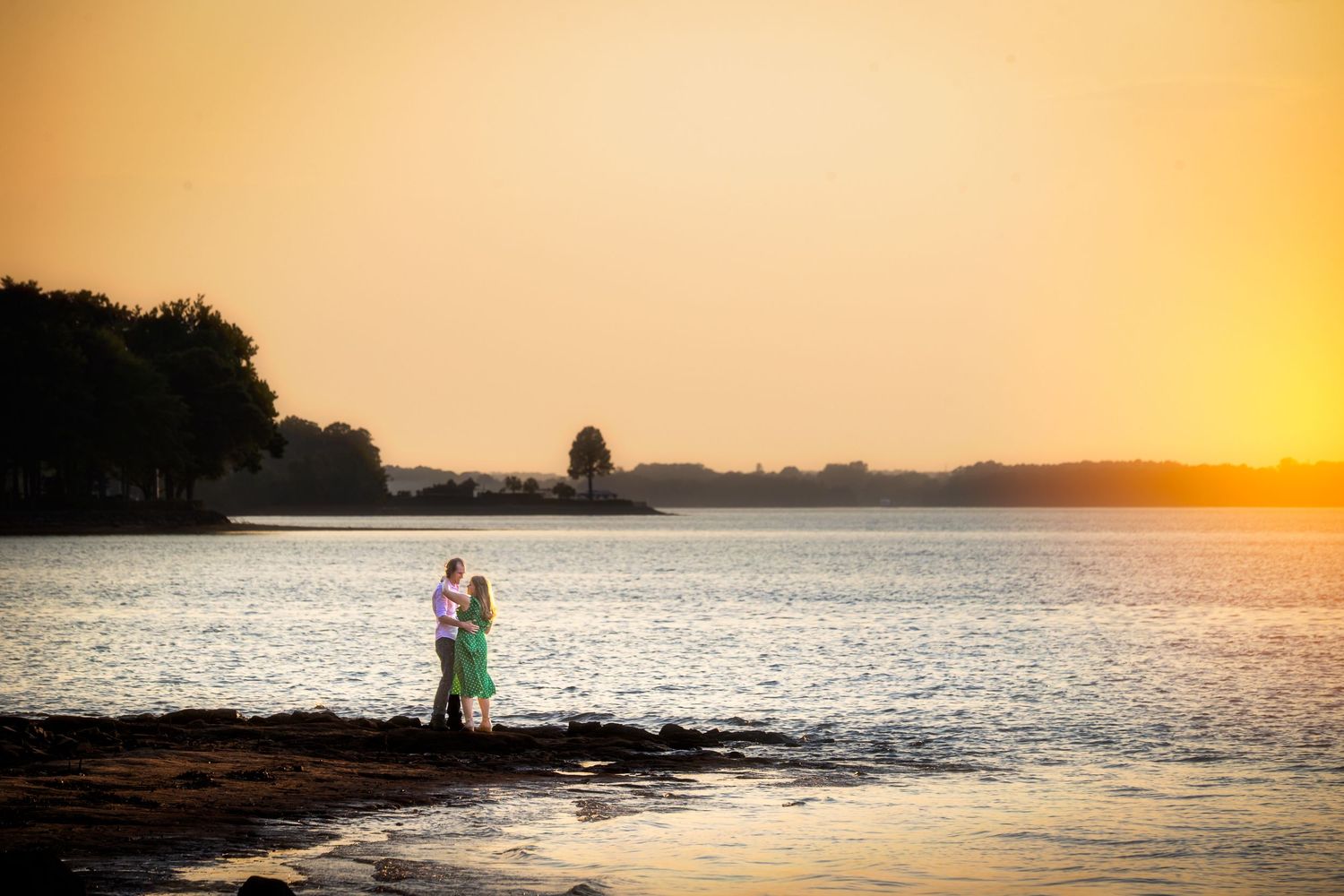 Couple embracing at sunset on rocky shoreline with golden sky reflecting on calm water.