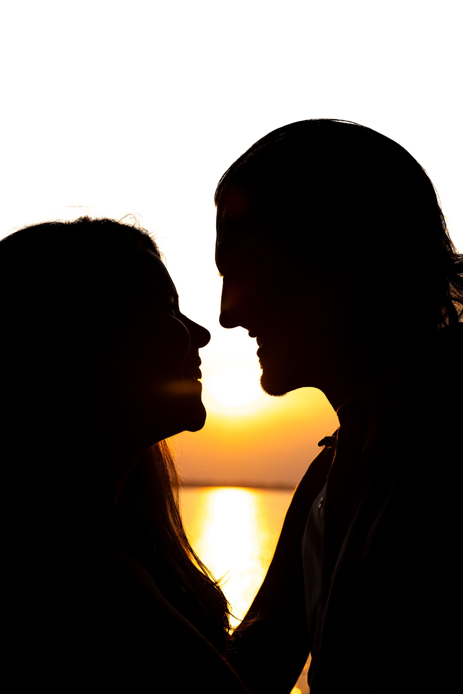 Romantic couple silhouette facing each other against a golden sunset over water.