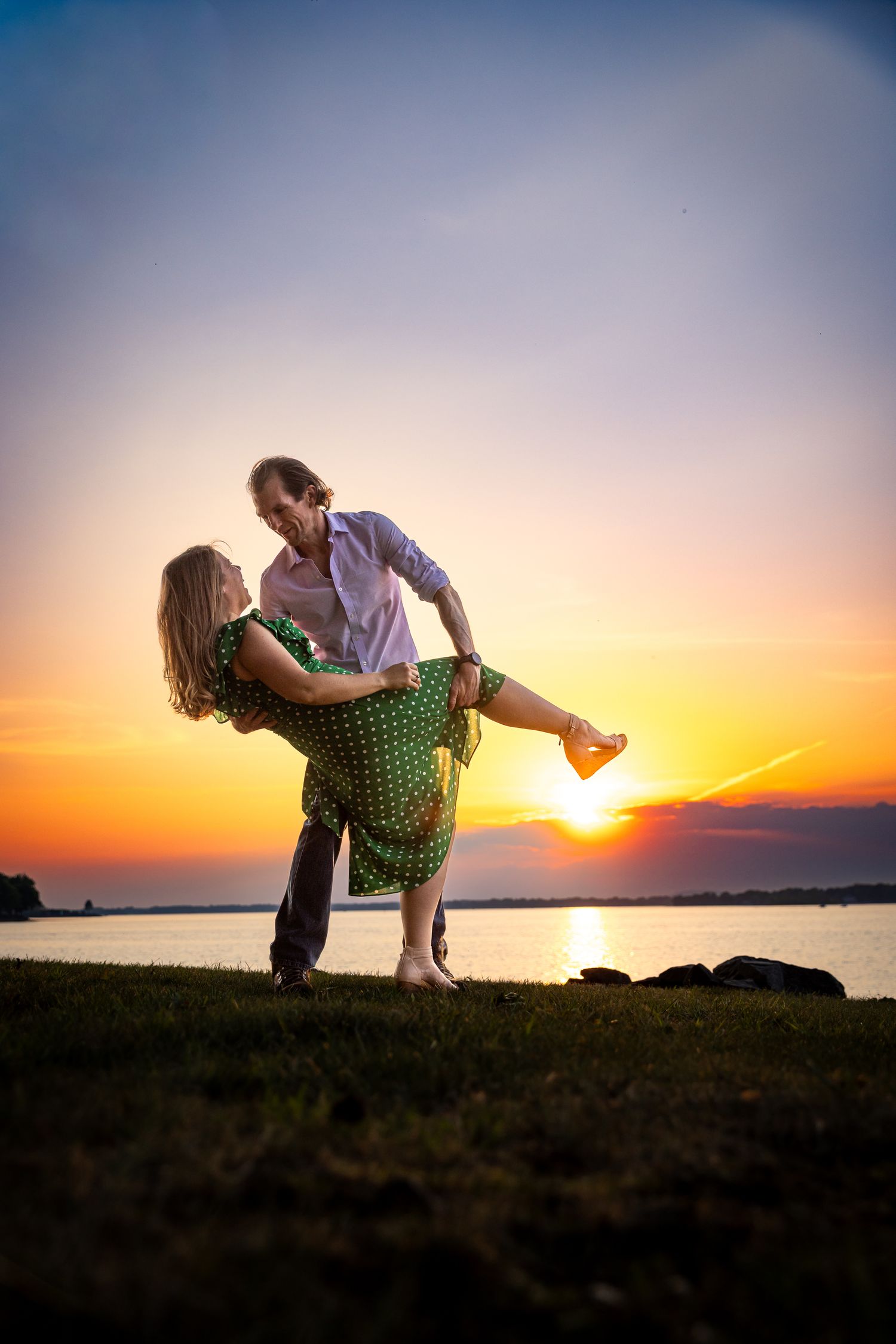 Couple dancing romantically at sunset by a lake, man lifting woman in green polka dot dress.