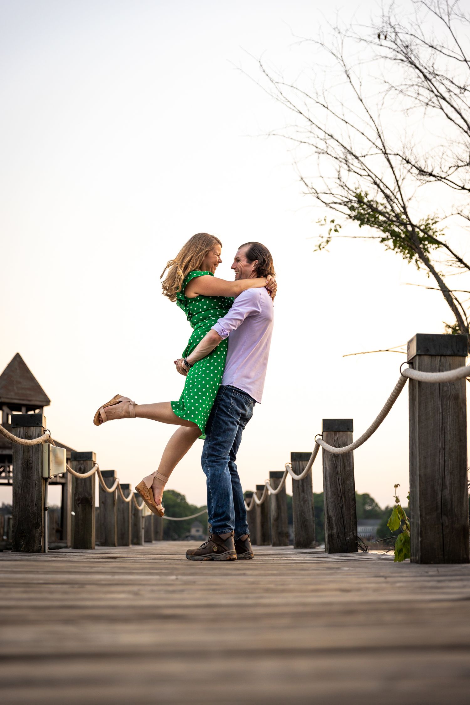 Man lifting woman in green dress on wooden pier at sunset.