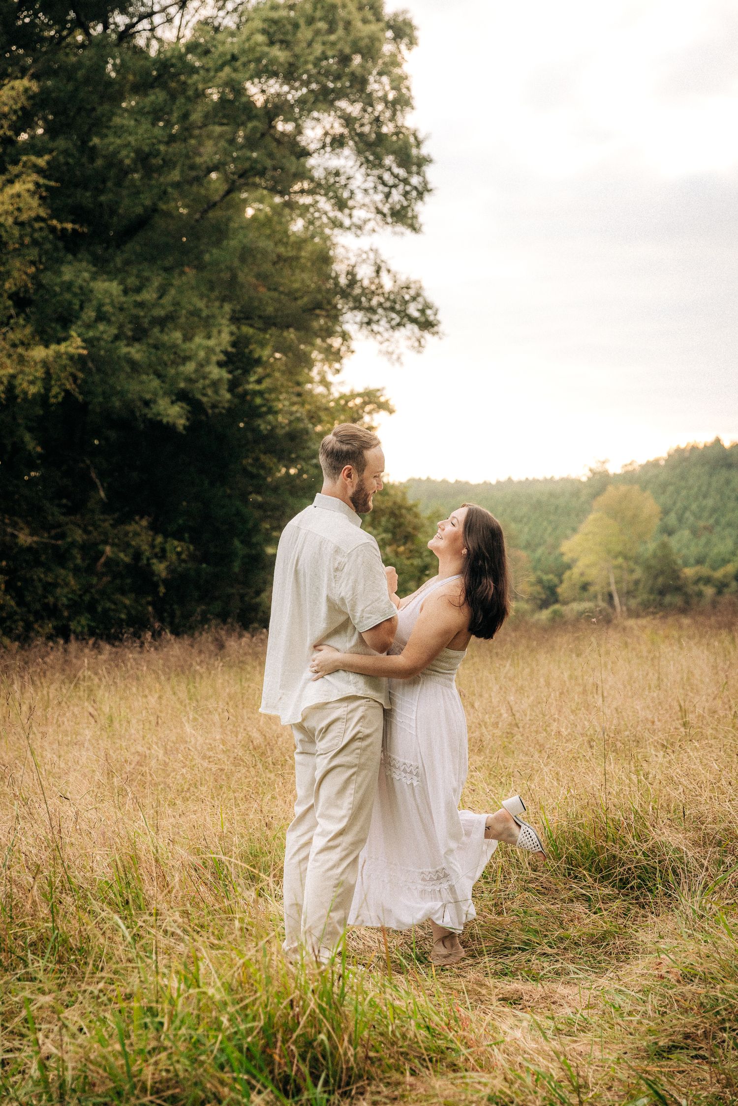 A couple in white outfits embrace in a sunlit meadow with trees in the background during golden hour.