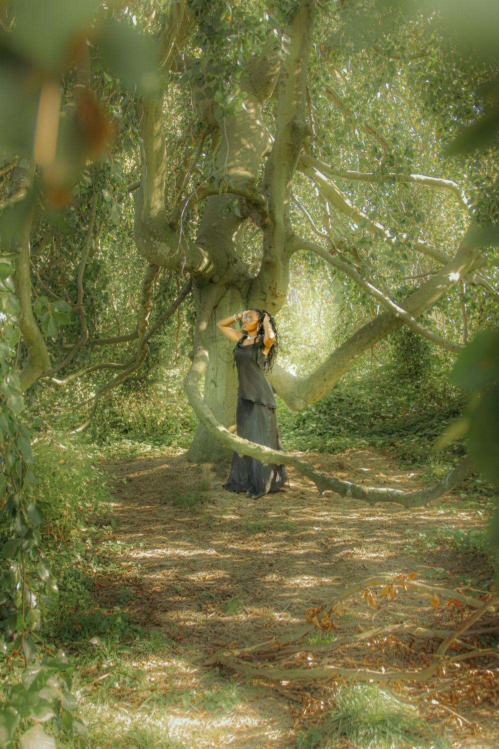 Portrait of a woman standing under a majestic tree in a long, modest dress, surrounded by nature exuding a dreamy and enchanting atmosphere.
