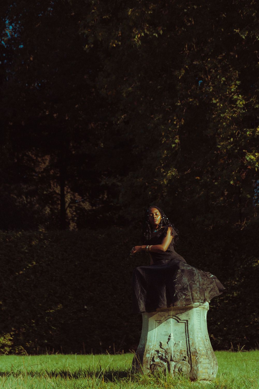 Model with closed eyes sitting on a weathered statue, immersed in nature, radiating tranquility.