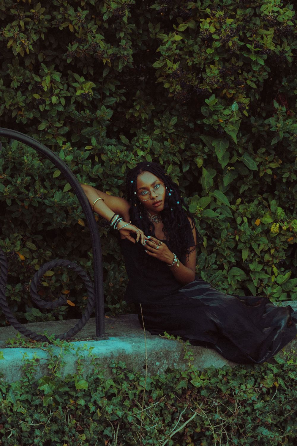 Model sitting on a stone bench, leaning against an architectural arch, surrounded by lush plants above and below.