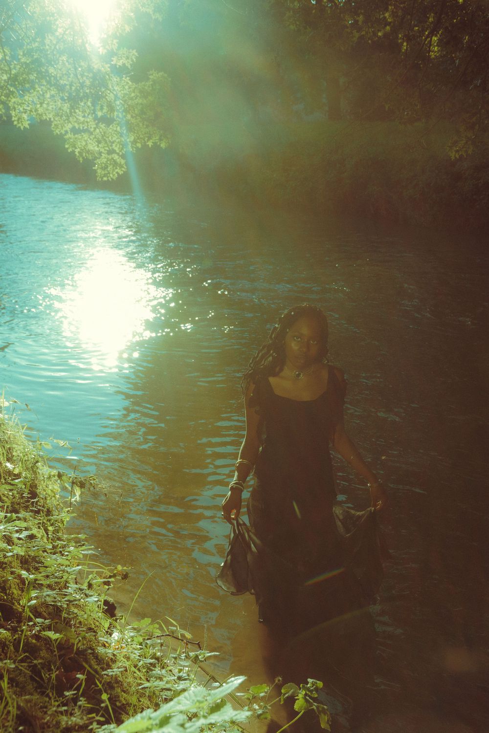 Woman in a flowing black dress standing in a river in Leuven, with water reflections creating a mystical, otherworldly atmosphere.