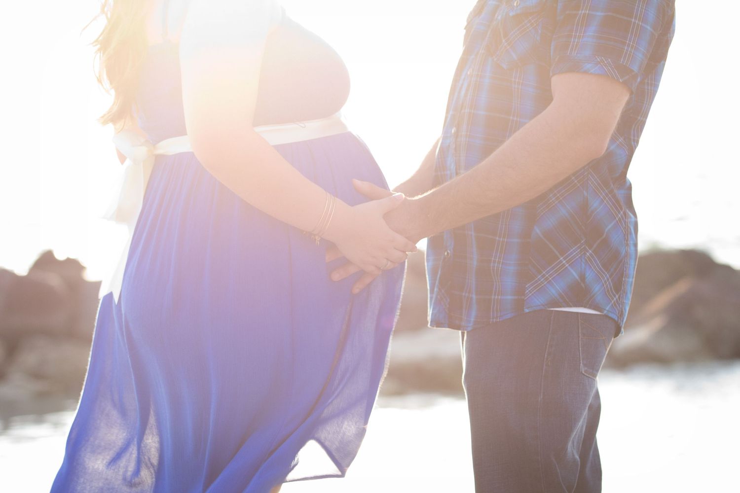 Sunlit couple holds hands over a pregnant belly, with the woman in a flowing blue dress and the man in a plaid shirt by the water.