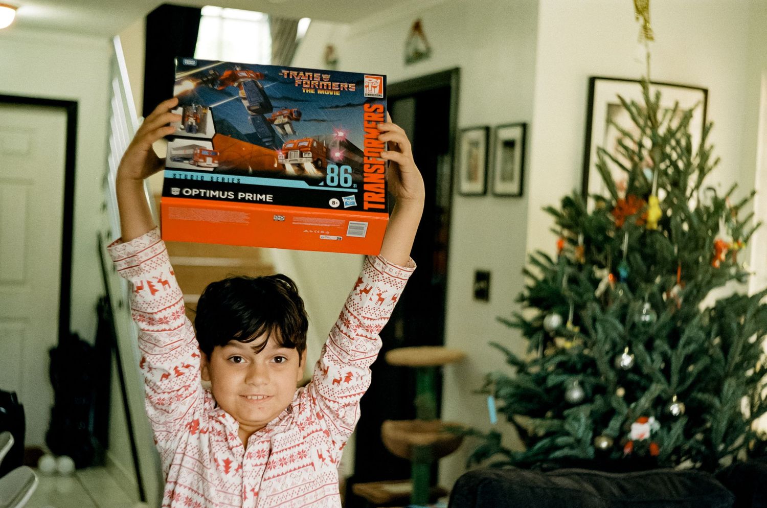 Child holding up a box in front of Christmas tree with excited expression.