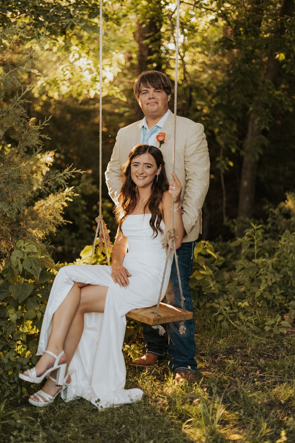 Romantic couple sitting on wooden swing in sunlit forest during outdoor wedding photoshoot.
