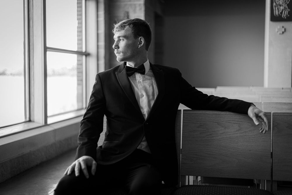 A person in a black suit gazes out a window in a dramatic black and white photograph.