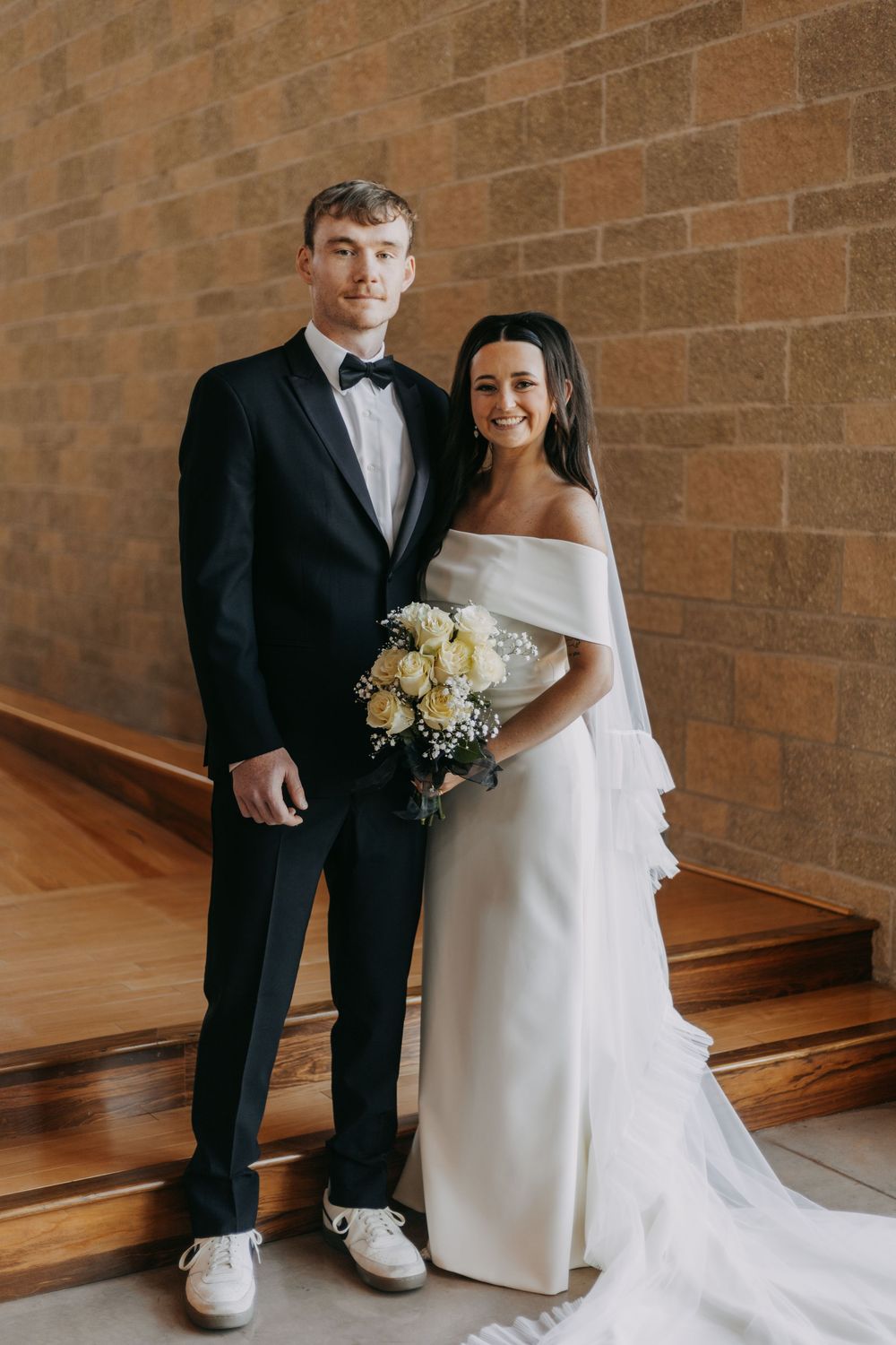 A bride and groom pose together in front of a brick wall wearing traditional wedding attire with white flowers.