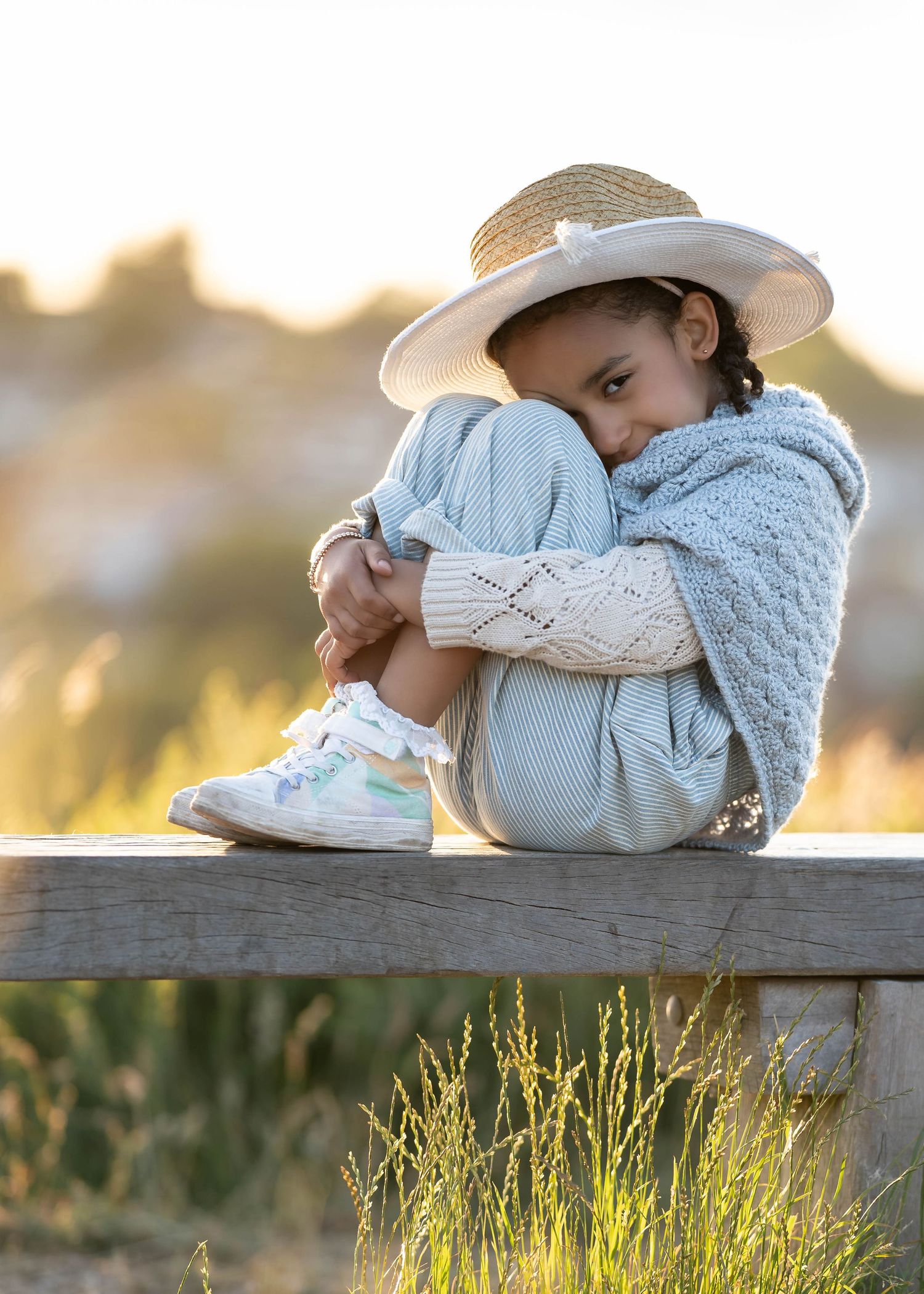 Child in a sun hat and sweater sitting on wooden bench during golden hour.