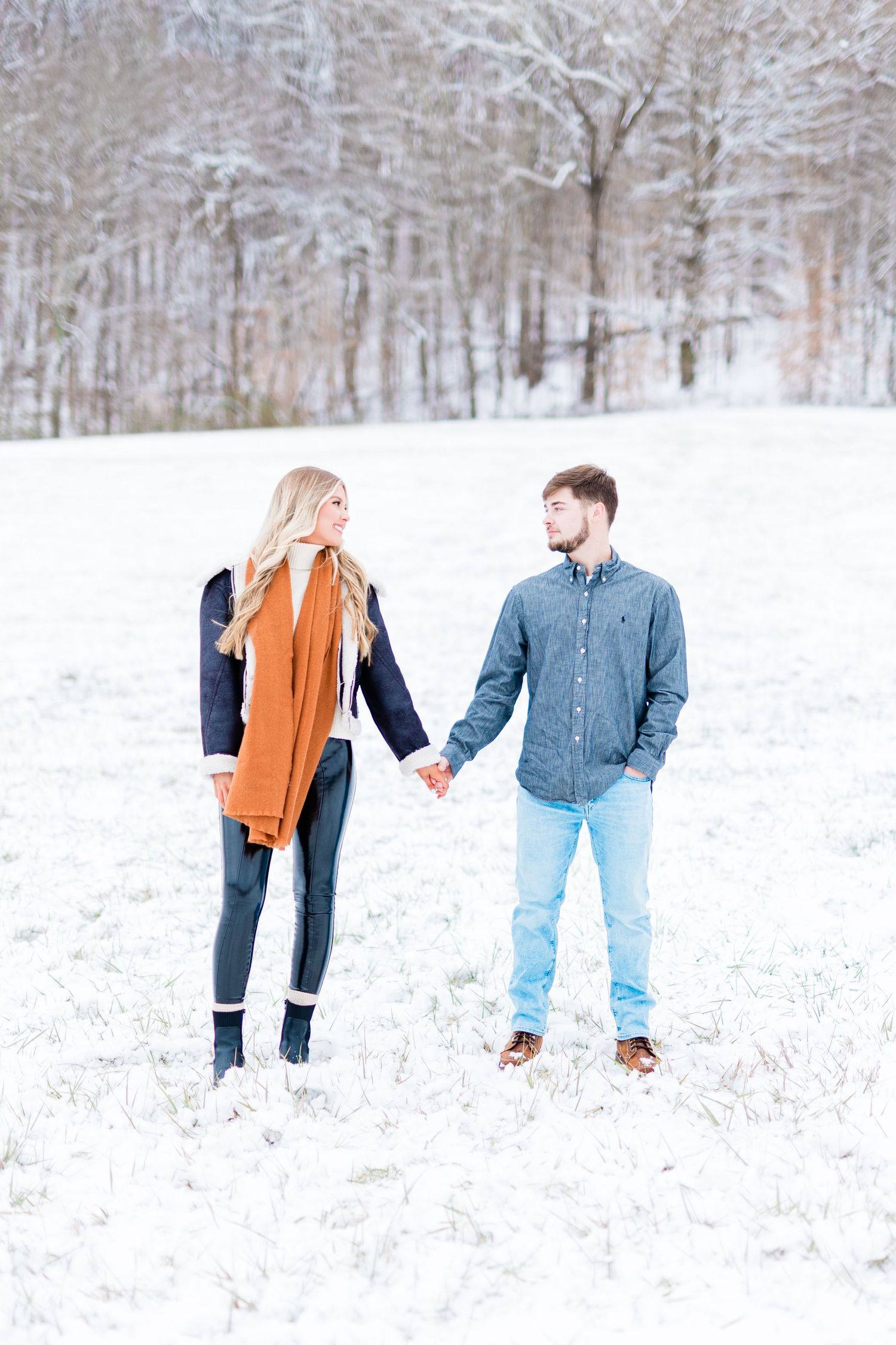 A couple holding hands while standing in a snowy winter landscape with bare trees in background.
