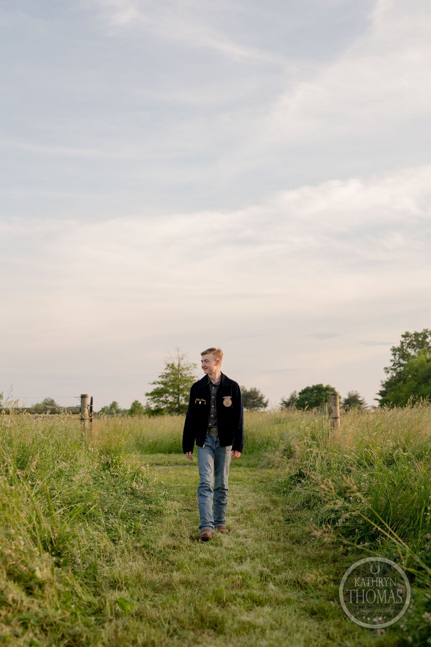 Student in FFA jacket walks along grassy path between fence posts.
