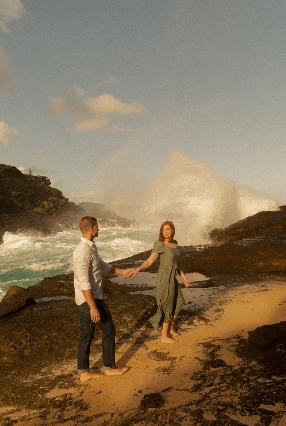 A couple stands on rocky coastline with crashing waves and dramatic sunset lighting.