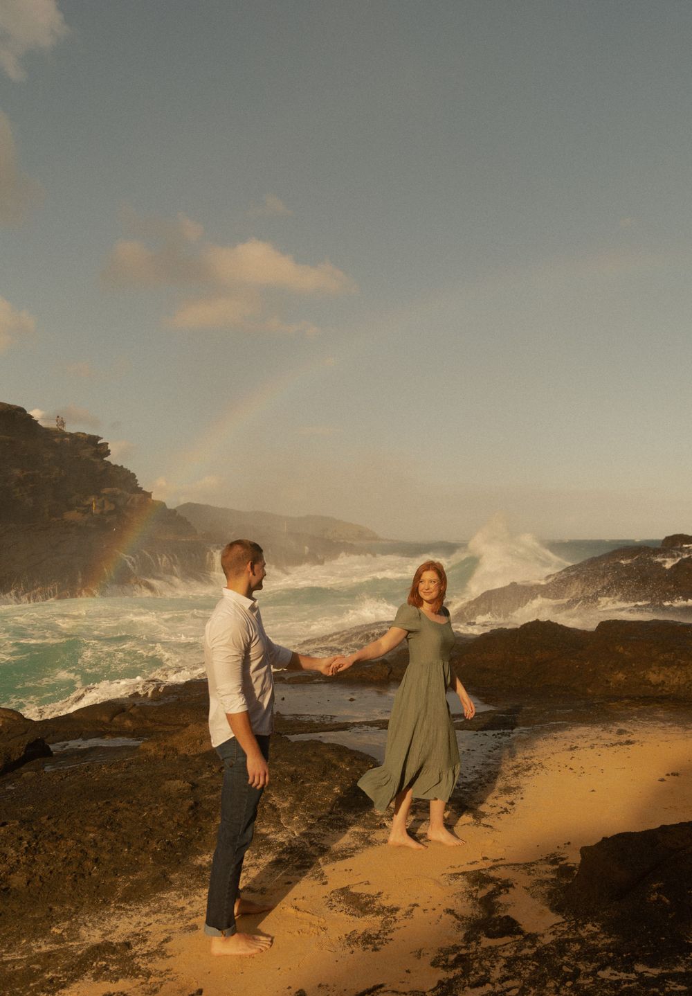 A couple holding hands on a dramatic coastline with waves crashing behind them.