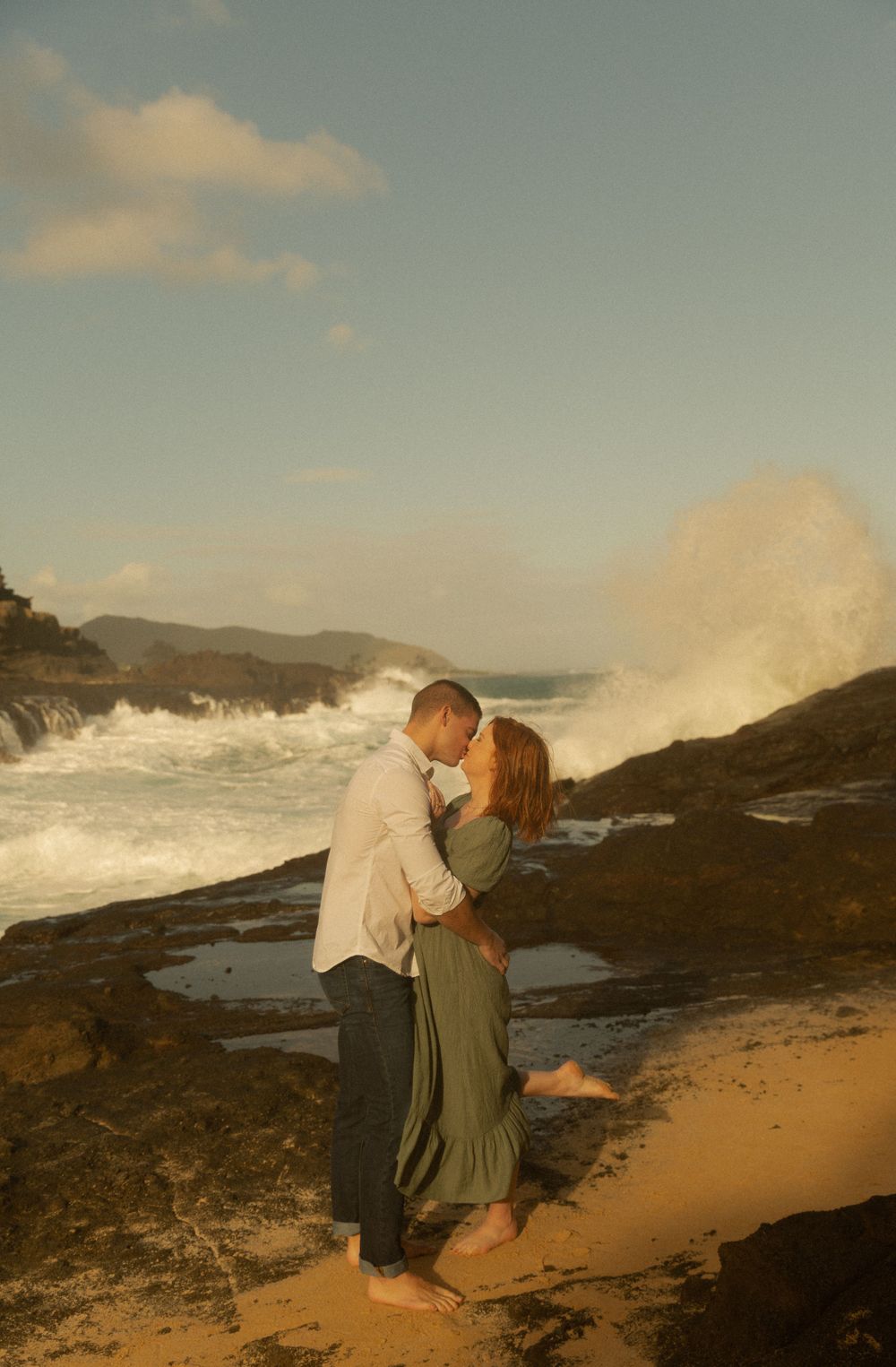 A romantic embrace on a rocky beach with ocean waves crashing in the background.