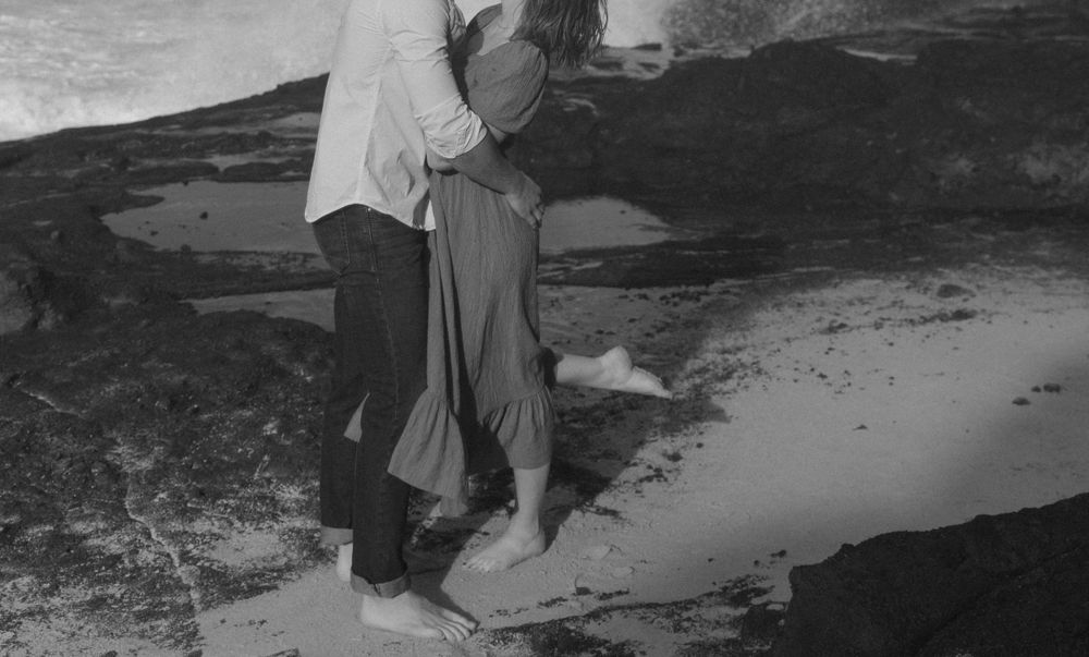 A black and white photo of a couple embracing on rocky coastal terrain.