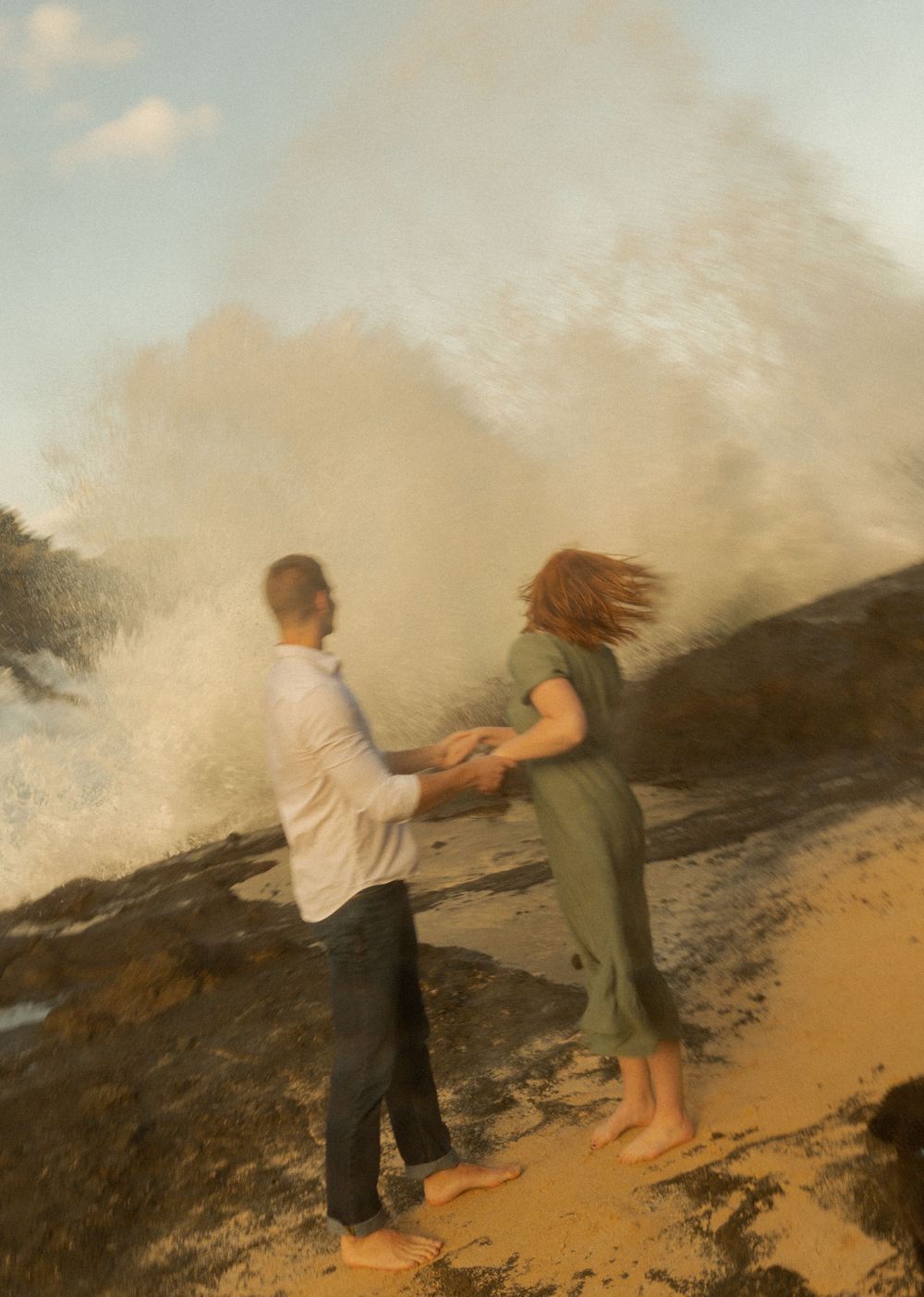 A couple holding hands as ocean spray dramatically erupts behind them on a rocky shore.