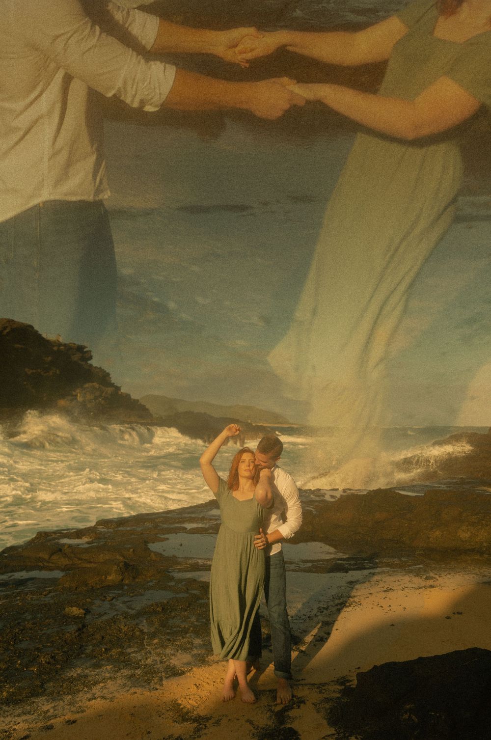 A couple embraces on a rocky coastline as waves crash against the shore at sunset.