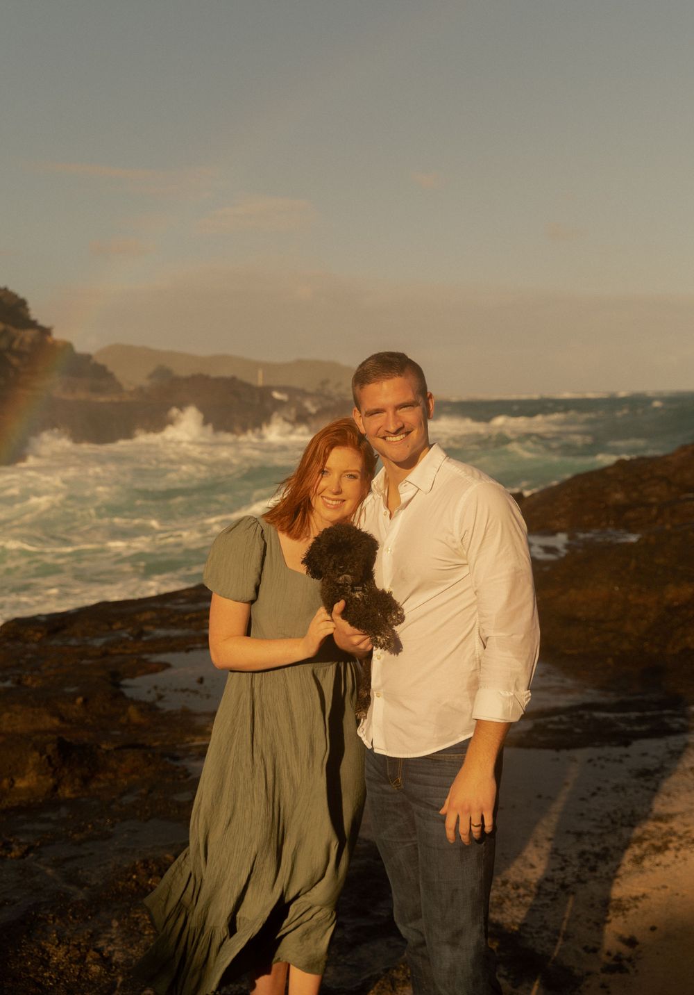 A couple holding a small black dog poses together on coastal rocks with crashing waves behind them.